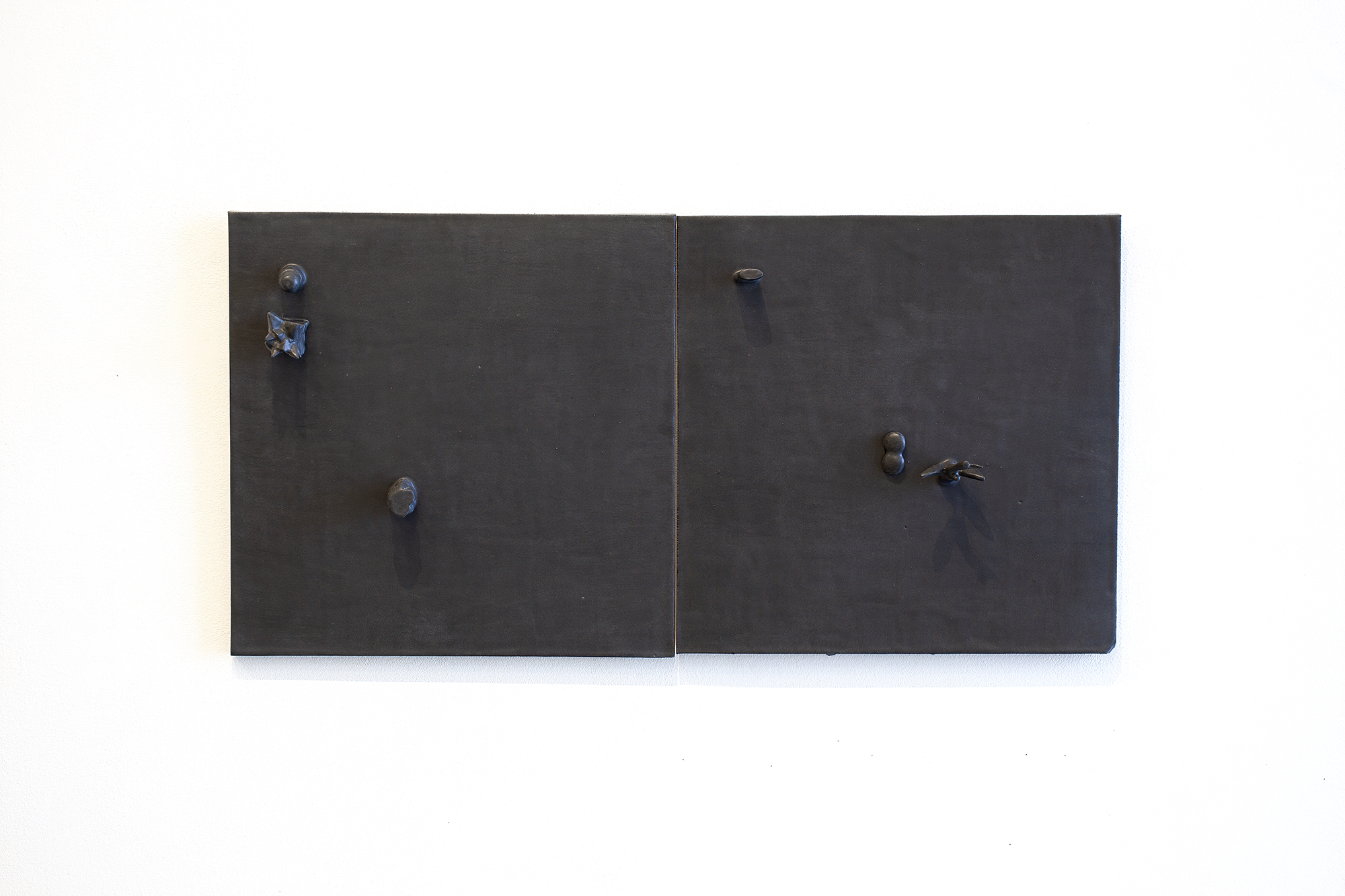 Two dark matte ceramic panels mounted side by side on a white wall, each fitted with metallic knobs and hardware. The left panel features two circular knobs positioned vertically, while the right panel has a similar knob with an additional key or lever mechanism, creating an austere, minimalist composition suggestive of functional cabinetry.