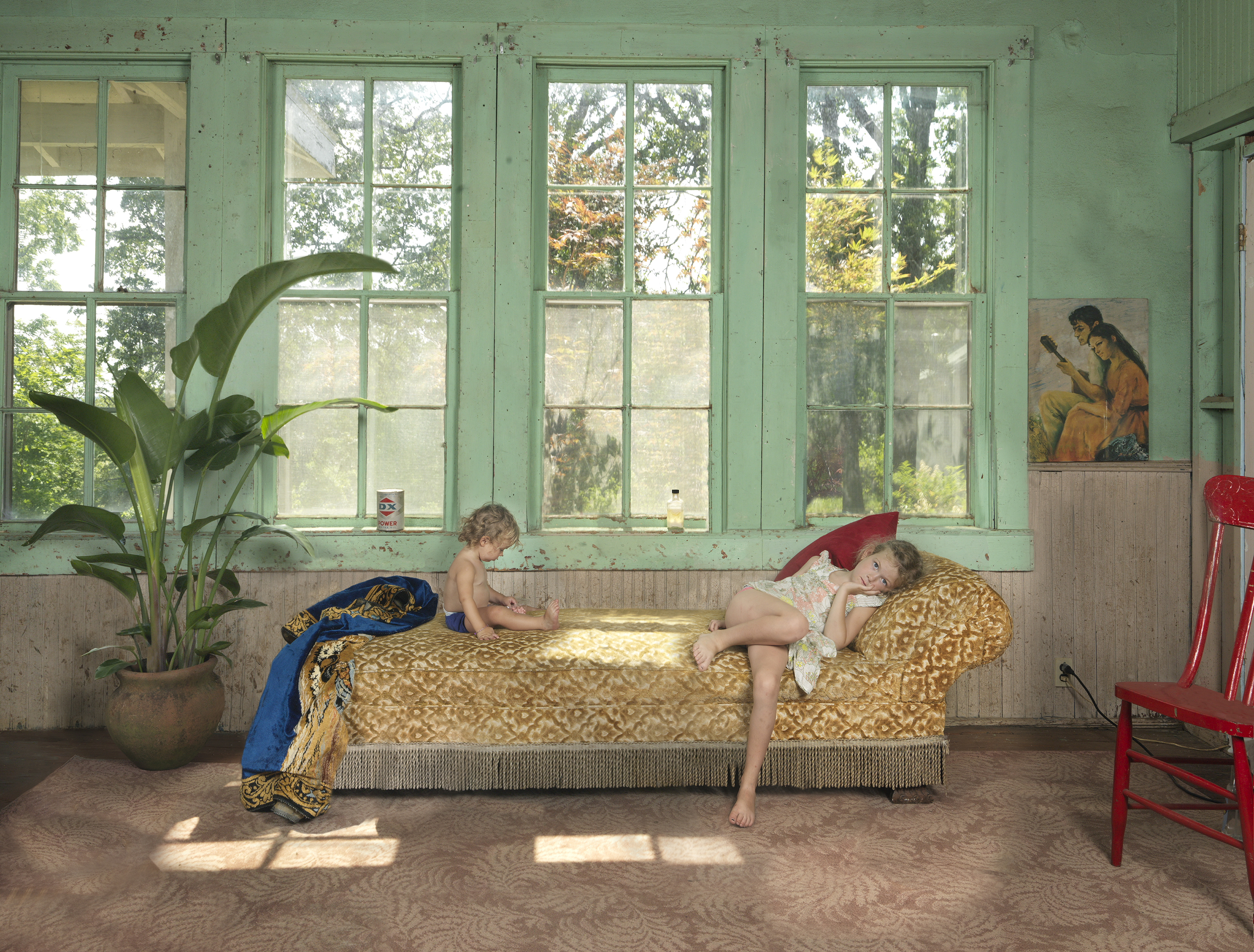 Two children play and rest on a patterned daybed in a sunlit room with mint-green walls and large gridded windows overlooking trees. A potted plant, red chair, and framed artwork are visible in the softly lit interior space.