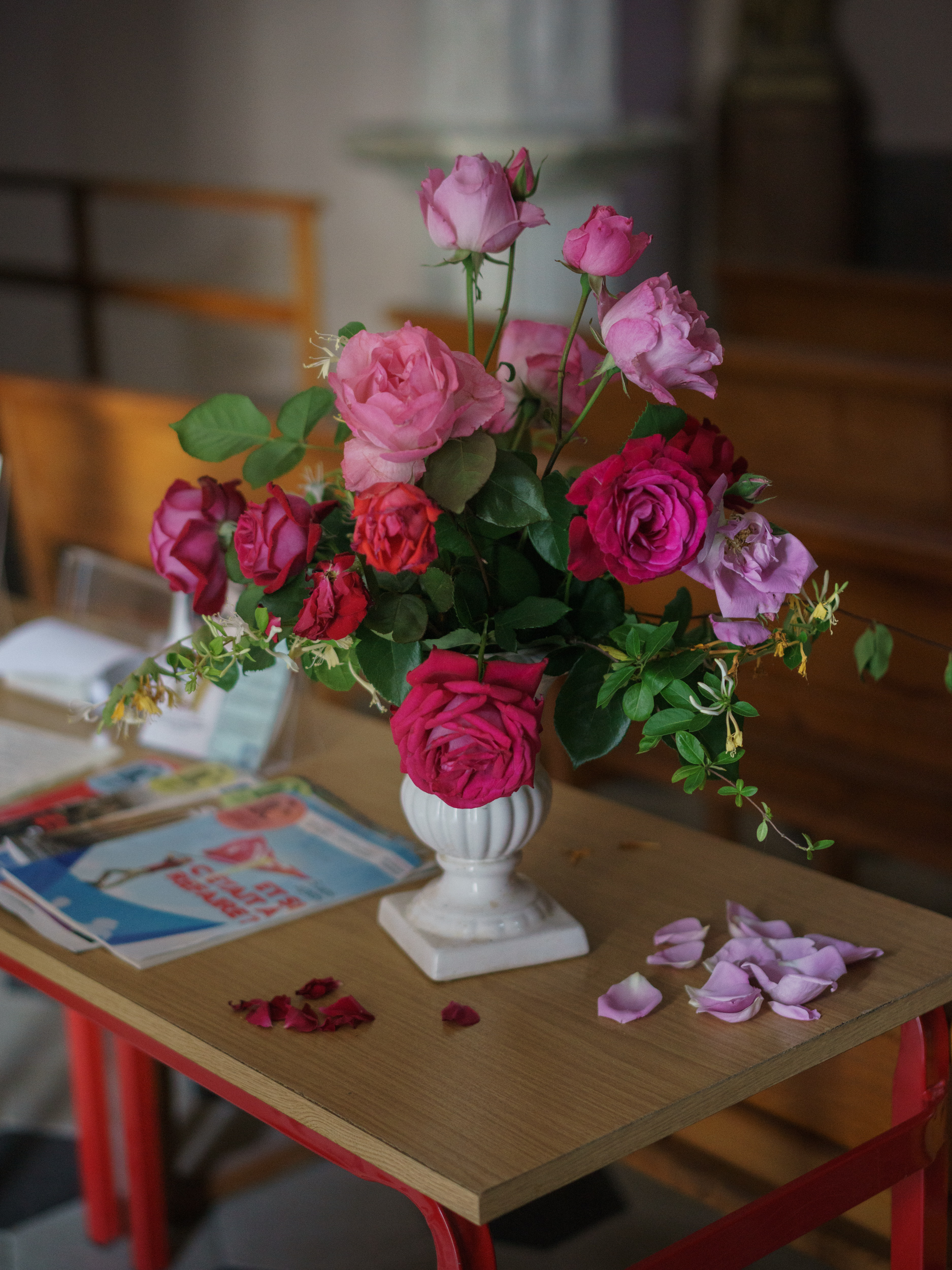 A vibrant floral arrangement of pink, magenta, and lavender roses in various stages of bloom sits in a white pedestal vase on a wooden table with red legs. Scattered rose petals in deep crimson and pale purple rest on the table surface beside the arrangement, with magazines and indoor furnishings visible softly blurred in the background.