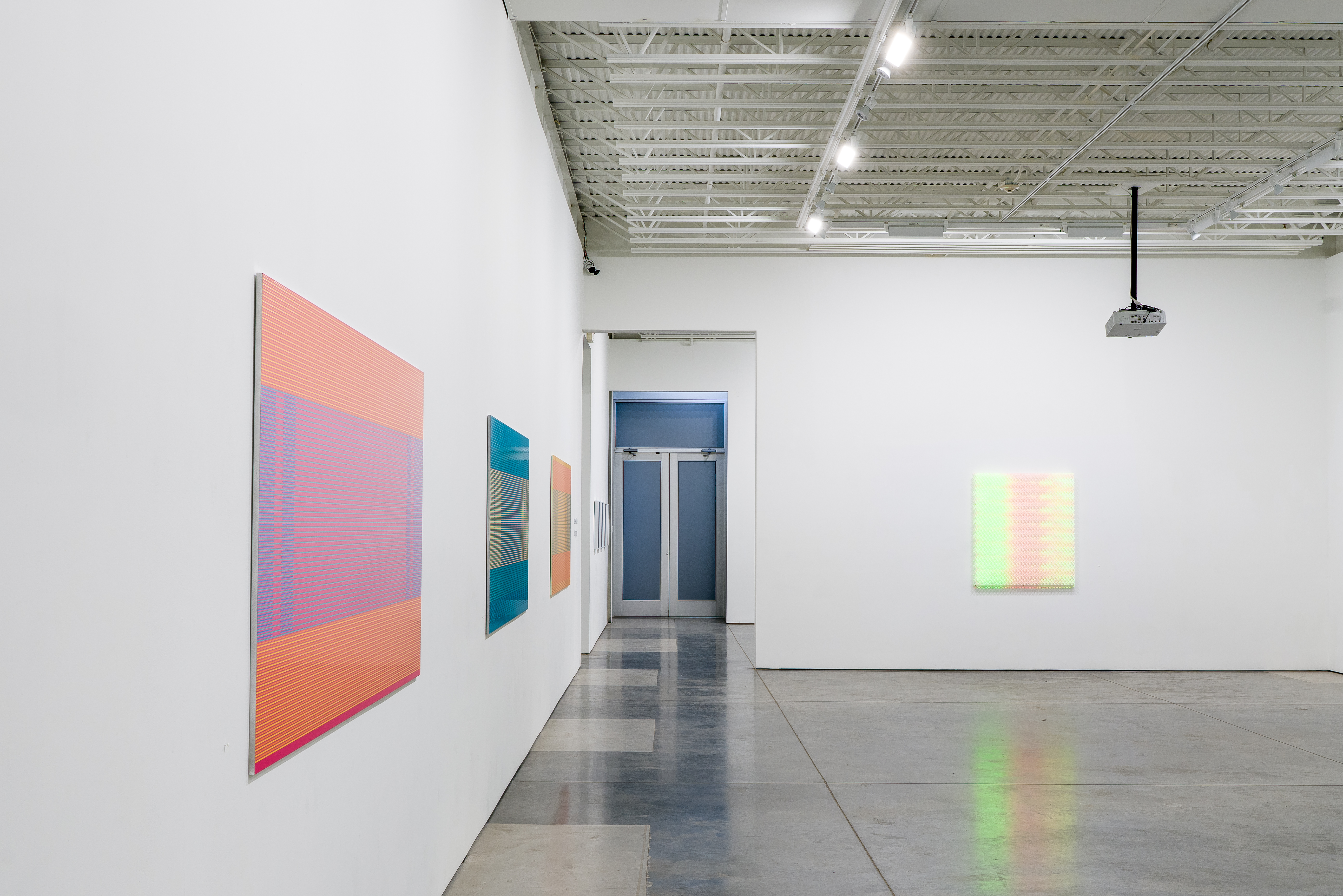 Gallery installation featuring four large-scale works combining painting and digital projection. Left wall displays a vertical composition with horizontal striations in coral, pink, and purple tones. Center works include a teal and orange geometric piece and a smaller tan panel. Right wall shows a luminous composition with soft green and peachy gradients. Polished concrete floor reflects the projected light. Industrial ceiling with exposed beams and track lighting. White gallery walls and blue doorway visible in background.