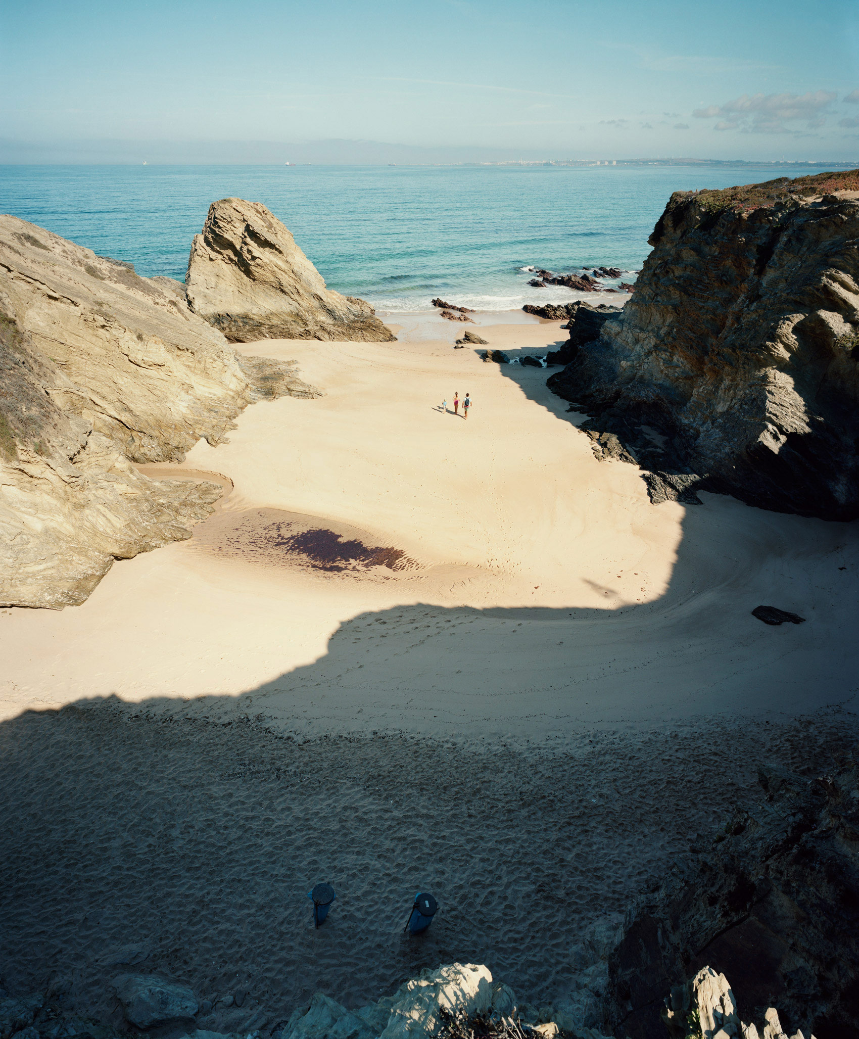 A secluded beach cove framed by dramatic pale limestone cliffs and dark rocky outcroppings, with turquoise water beyond and a few figures visible on the pristine sand. Shadows cast by the rocks create strong geometric patterns across the foreground and beach.