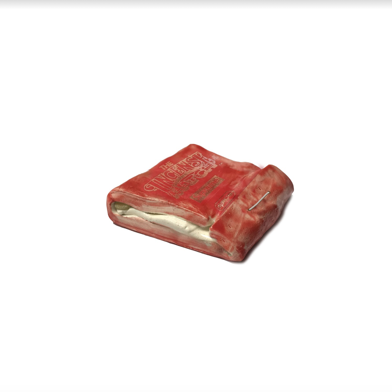 A red ceramic object resembling a worn matchbook, with embossed text visible on its surface and cream-colored interior pages visible where it opens slightly. The piece is glazed with a matte finish and accented with gold luster detailing.