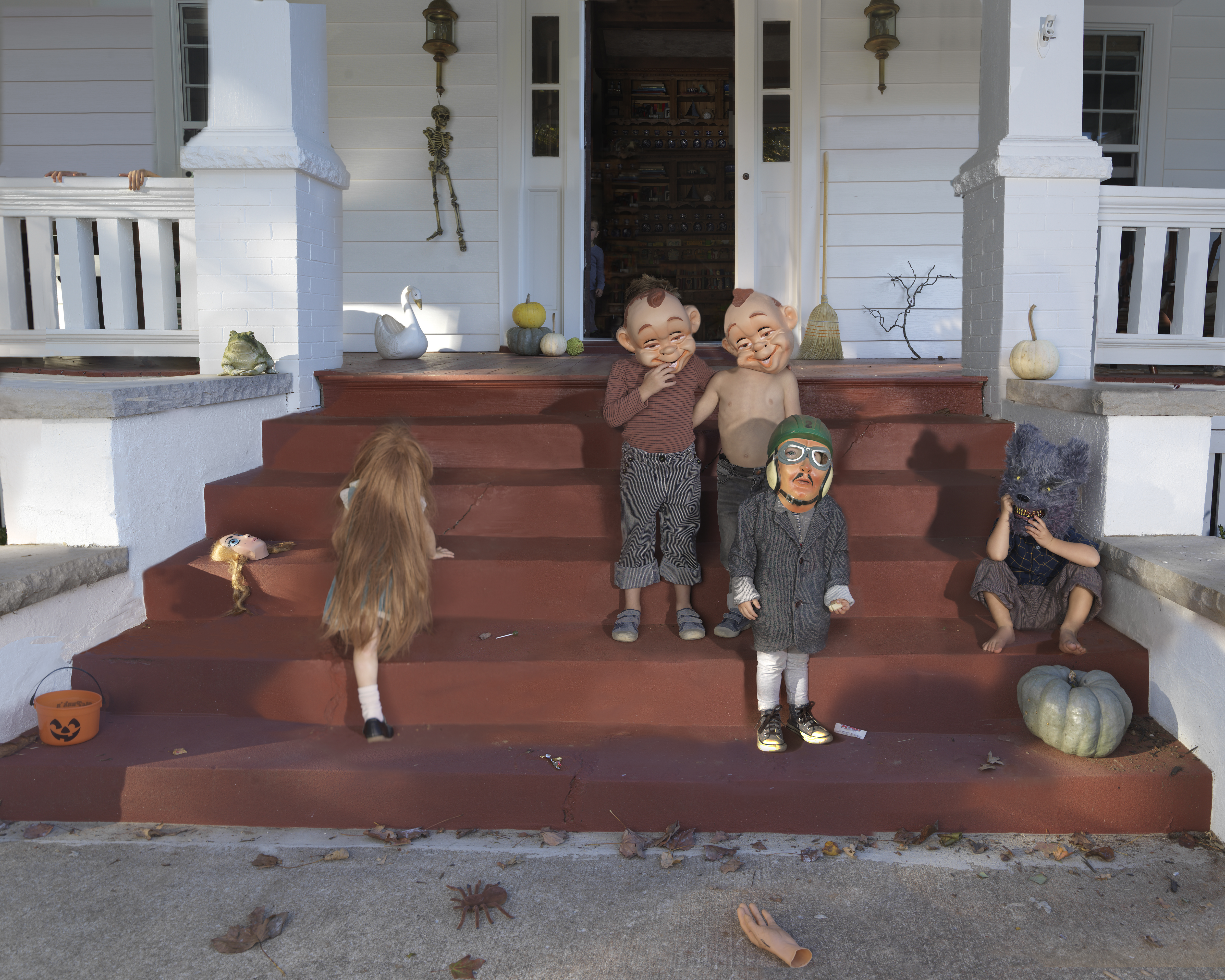 A surreal domestic scene featuring life-sized gnome and troll figurines arranged on brick front steps alongside real children in Halloween costumes. The white colonial-style house backdrop includes decorative gourds, hanging lanterns, and autumn foliage, creating an unsettling blend of the whimsical and uncanny through scale disruption and the intermingling of handmade figures with human figures.