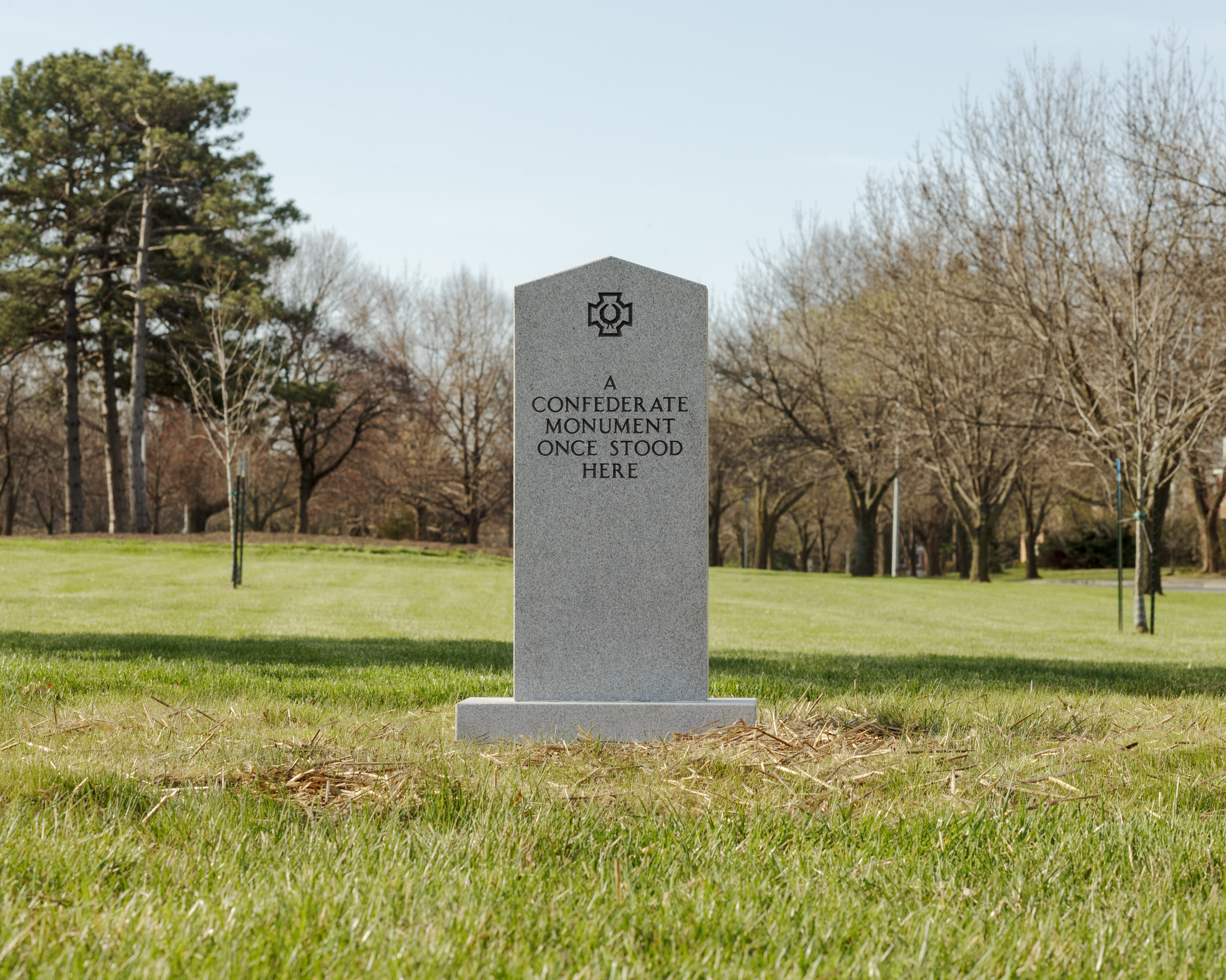 A gray stone monument stands alone in a grassy park surrounded by bare and evergreen trees. The obelisk-shaped marker bears an engraved symbol at its top and text reading "A CONFEDERATE MONUMENT ONCE STOOD HERE."