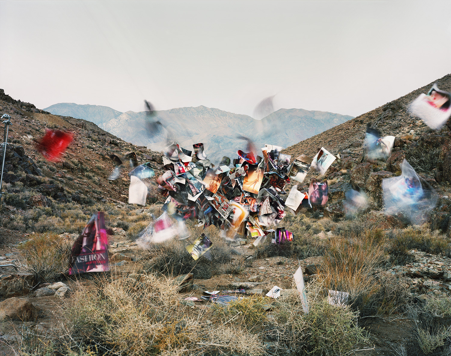 A dynamic collage of fashion magazines and colorful clothing spirals upward in a barren desert landscape, with mountains visible in the hazy background and sparse vegetation scattered across the rocky terrain below.