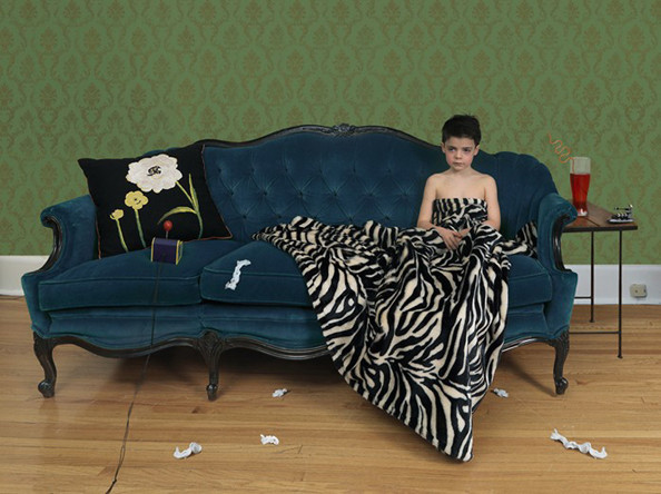 A person in a black strapless top reclines on a teal upholstered sofa draped with a zebra-striped blanket, against a green damask wallpapered wall. Scattered tissues and objects lie on the wooden floor, while a side table with a glass holds personal items nearby, creating a scene of domestic illness or malaise.
