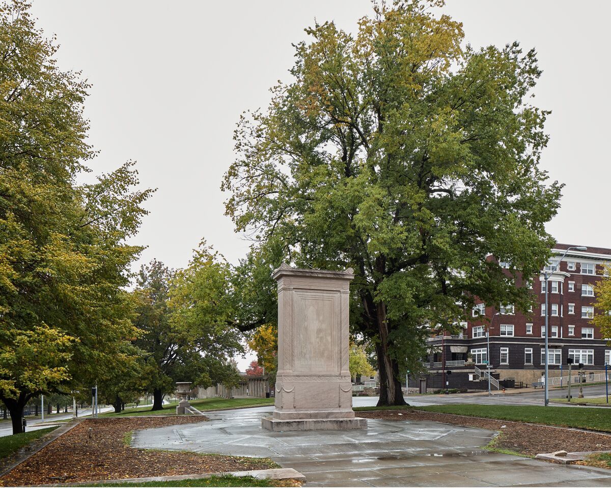 A wet stone monument pedestal stands centered in a public plaza, surrounded by mature trees in autumn foliage. Historic brick buildings frame the composition on the right, while tree-lined pathways extend into the distance on the left under an overcast sky.