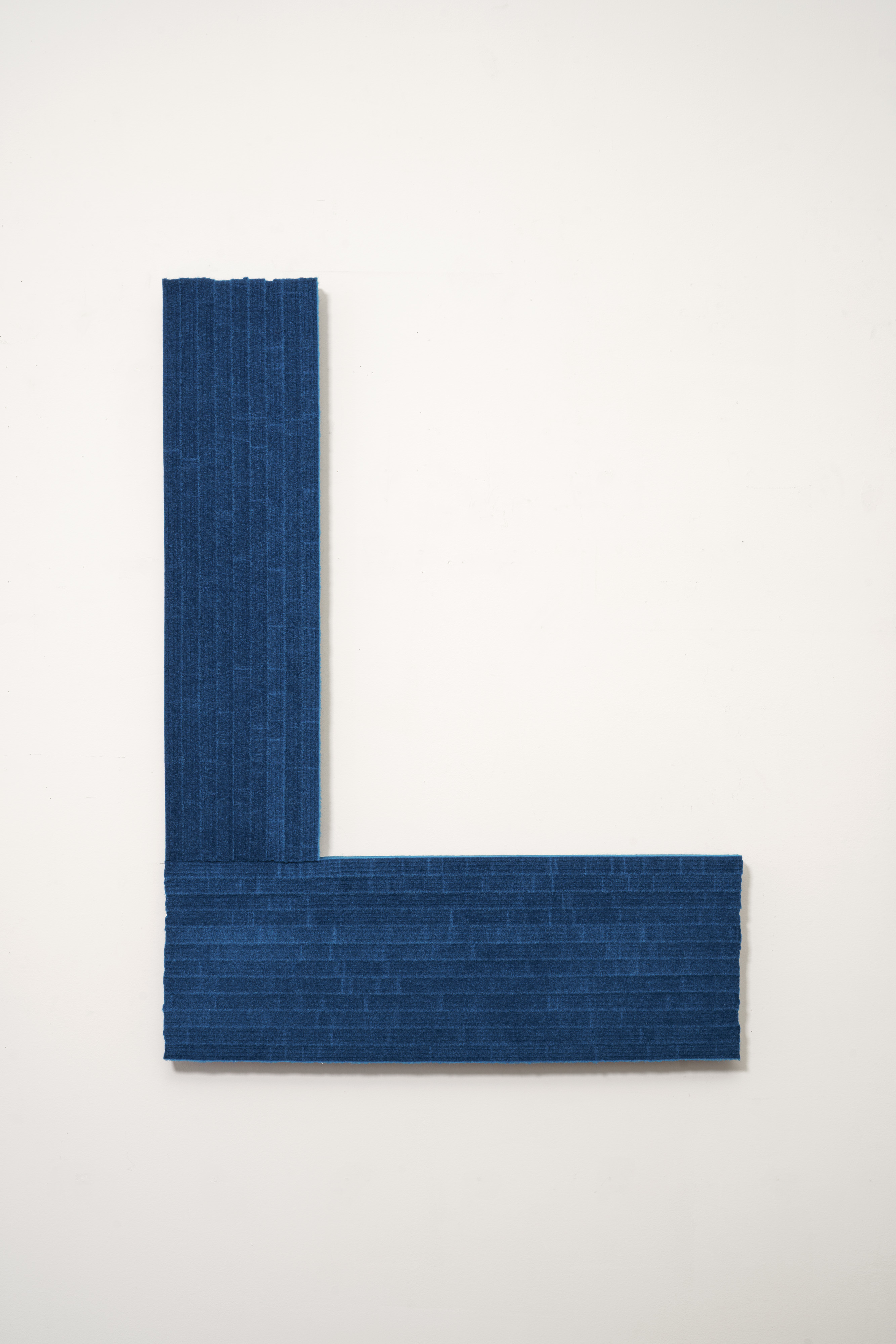 A deep blue textile panel arranged in an L-shaped composition against a white wall. The material has a subtle woven texture with visible vertical striations, creating a minimalist geometric form.