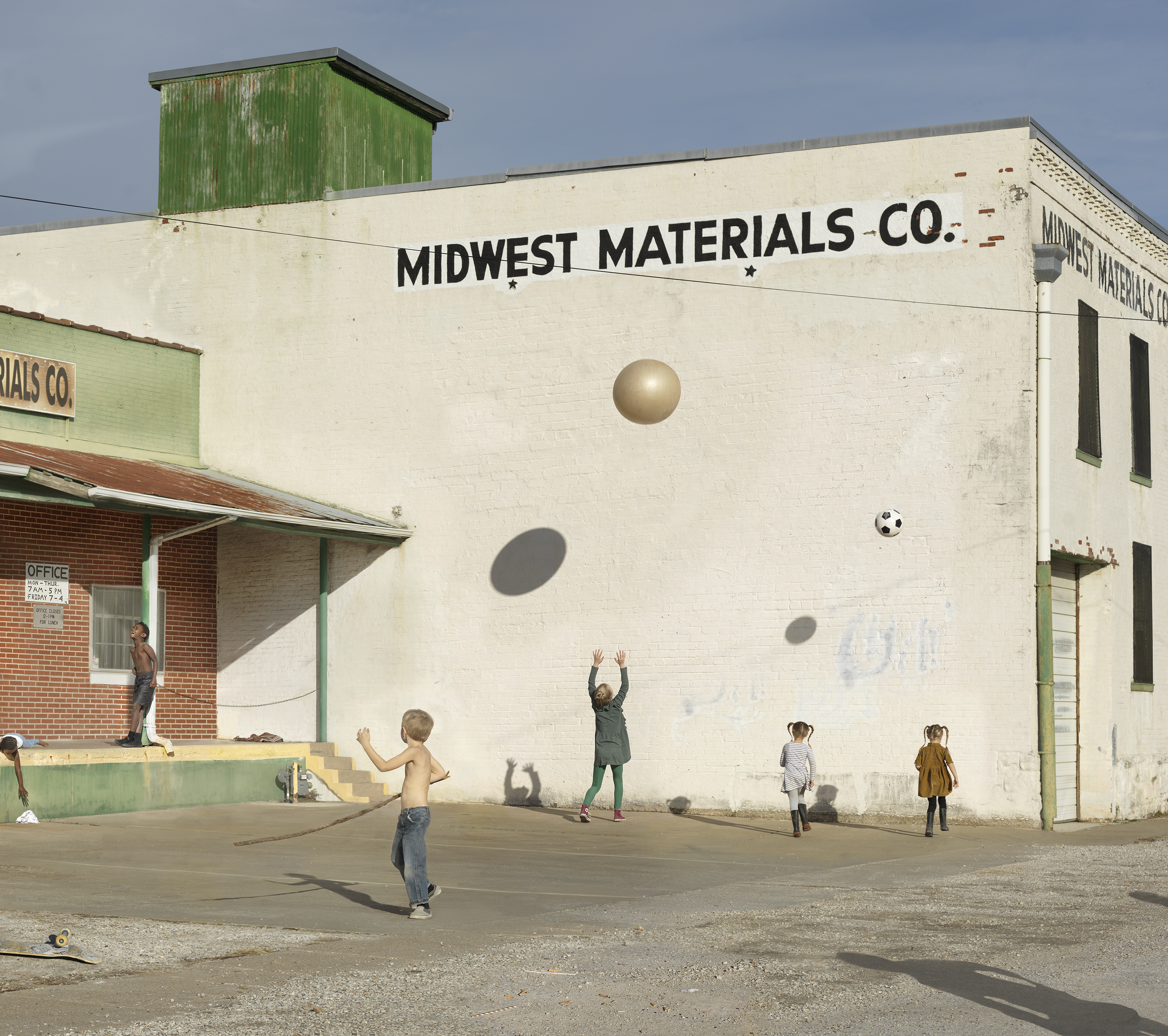 Children play in a sunny parking lot in front of a weathered cream-colored industrial building with "Midwest Materials Co." painted across its facade. Painted spheres and a soccer ball adorn the blank wall, transforming the utilitarian structure into an imaginative play space.
