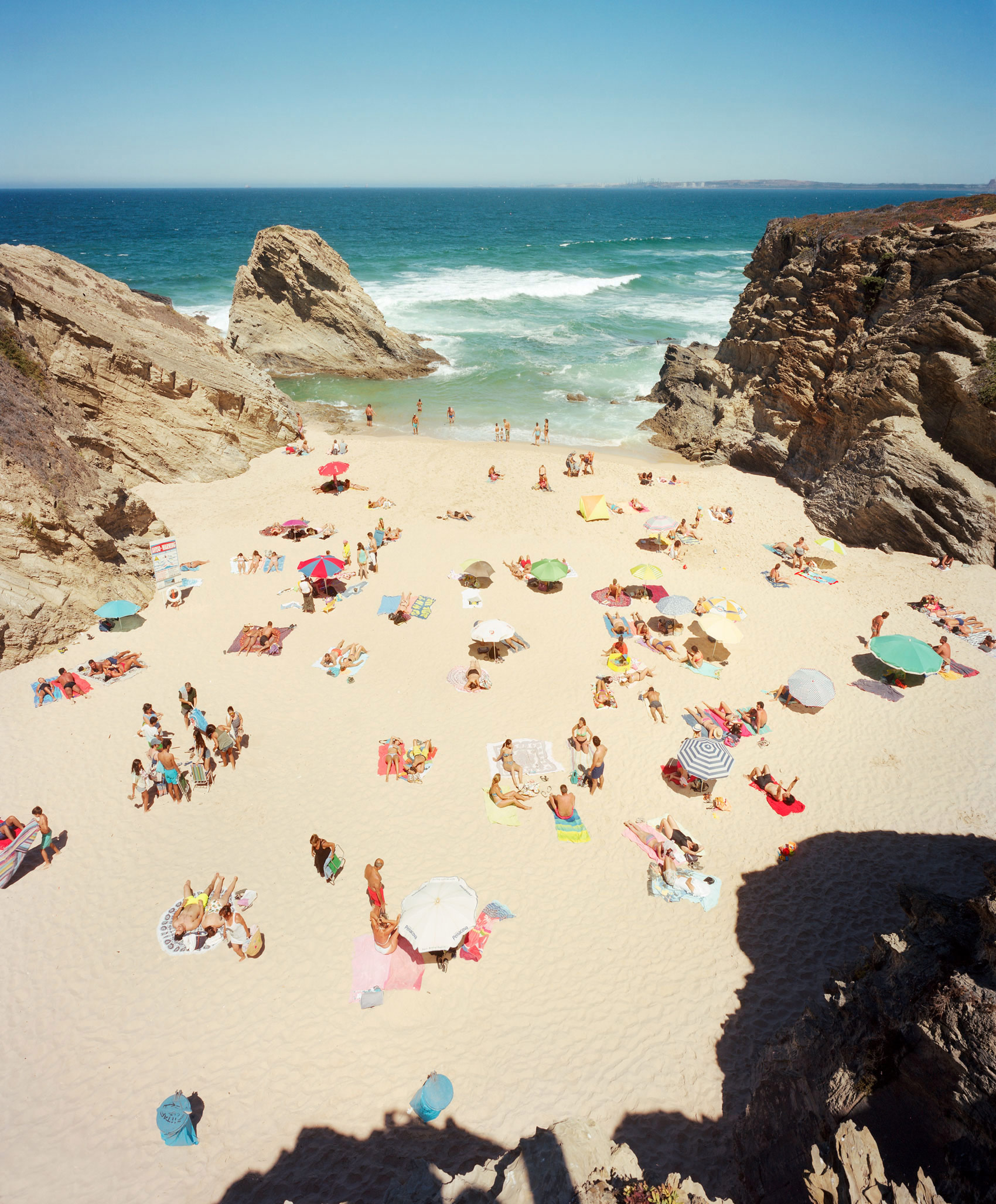 Aerial view of a crowded sandy beach cove enclosed by dramatic rocky cliffs, with beachgoers scattered across the sand beneath colorful umbrellas and towels, turquoise waves breaking along the shoreline, and a clear blue sky above.