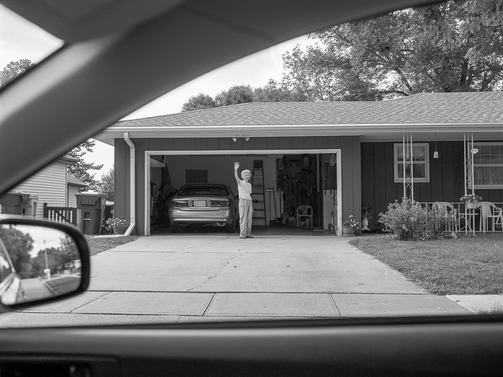 Black and white photograph taken from inside a car, framed by the windshield and side mirror, showing a woman in a white dress waving from the open garage of a modest single-story house with a parked sedan visible inside.
