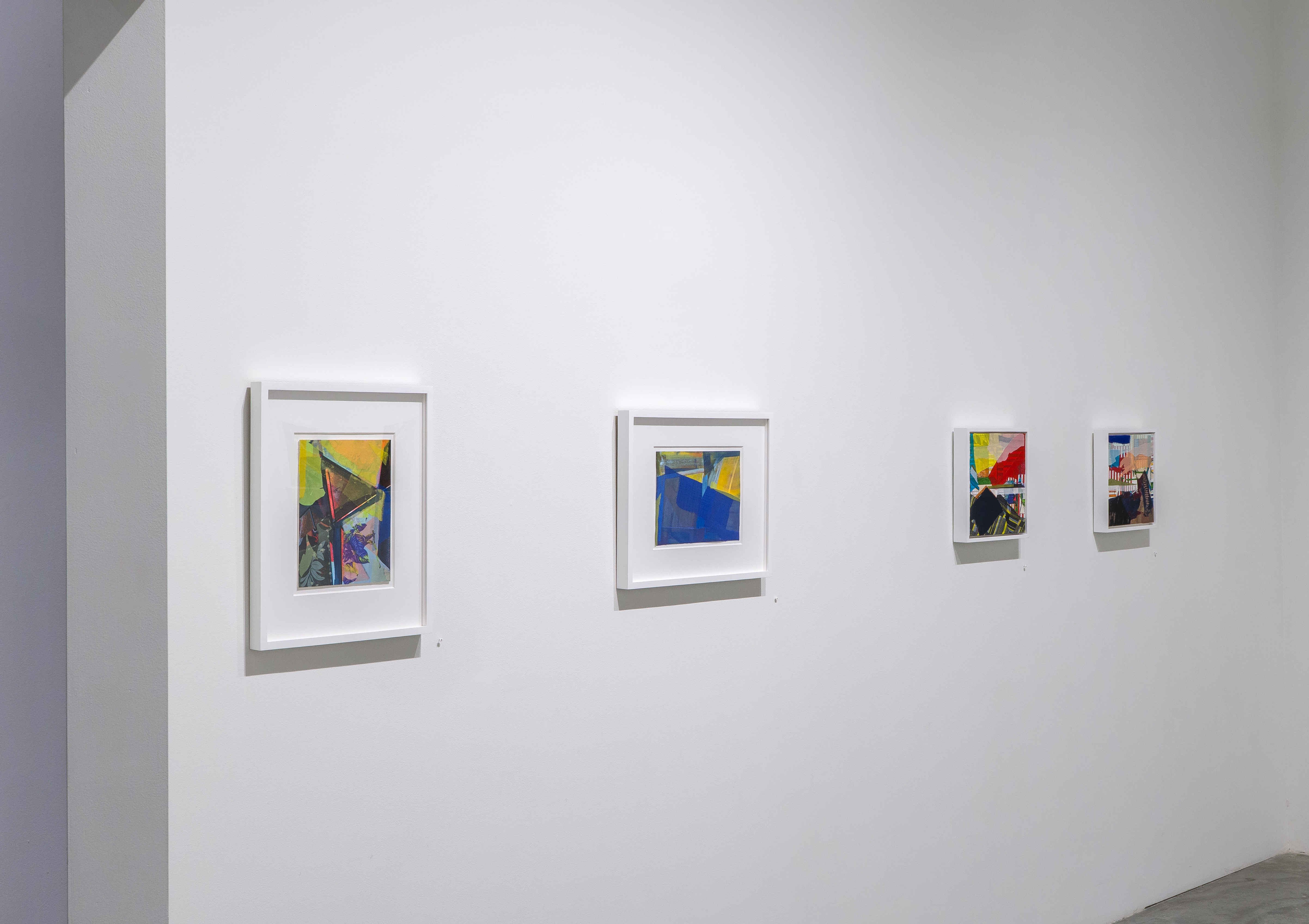 Four framed abstract works hang on a white gallery wall, displaying layered compositions in vibrant colors including yellows, blues, reds, and greens. The pieces combine mixed media elements with visible gestural marks and geometric forms, their frames creating shadow effects against the wall's clean surface.