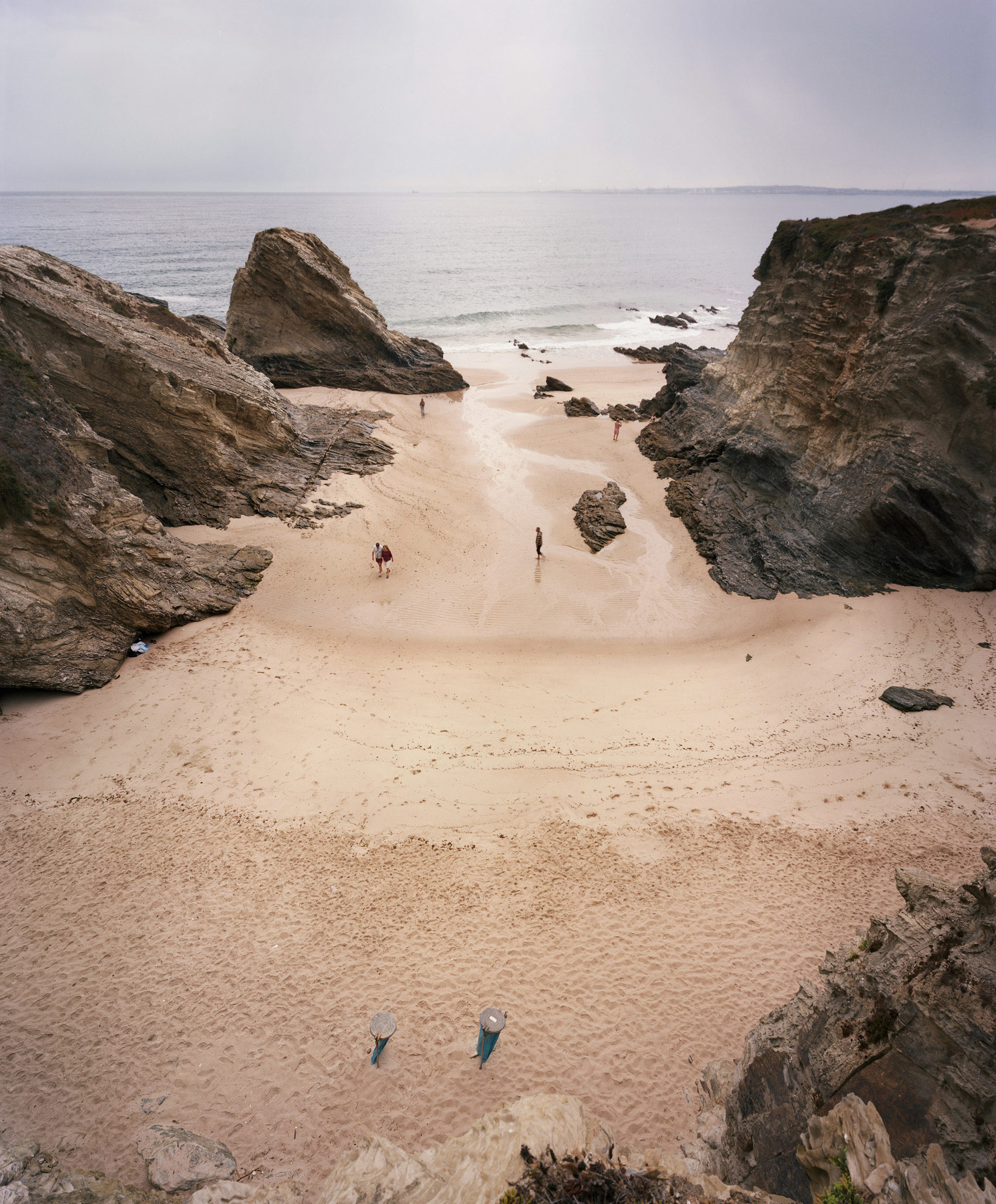 A secluded sandy beach enclosed by dramatic rocky outcrops and large boulders, with several small figures scattered across the sand and wading in shallow water. The composition is framed by weathered stone formations in the foreground and middle ground, while calm ocean and pale sky occupy the background.