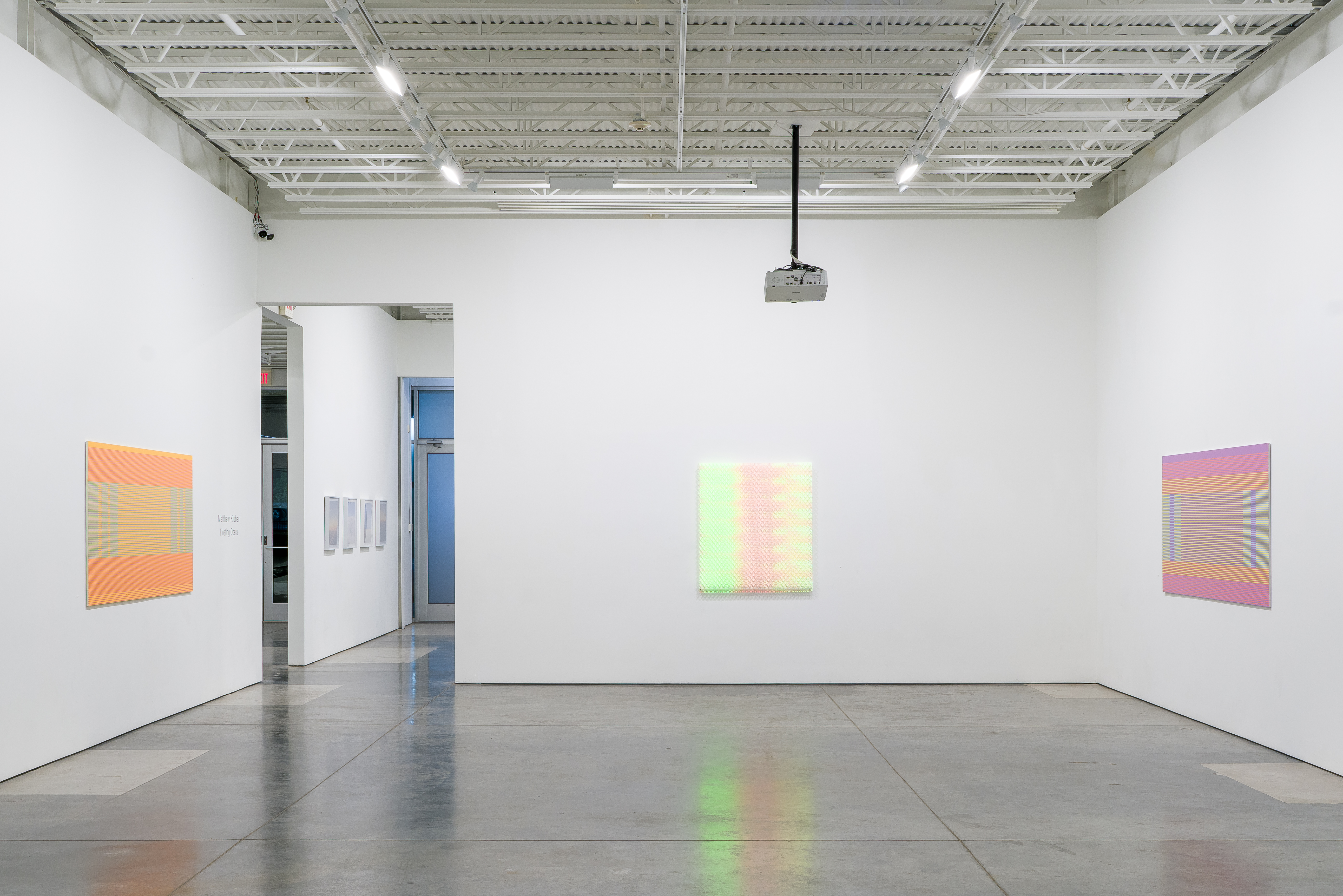 Contemporary gallery space with three illuminated artworks on white walls. Each piece features geometric patterns in vibrant colors—orange and yellow on the left, soft pastels in the center, and pink and purple on the right—created through the intersection of painted surfaces and projected digital light, casting colored reflections on the polished concrete floor.