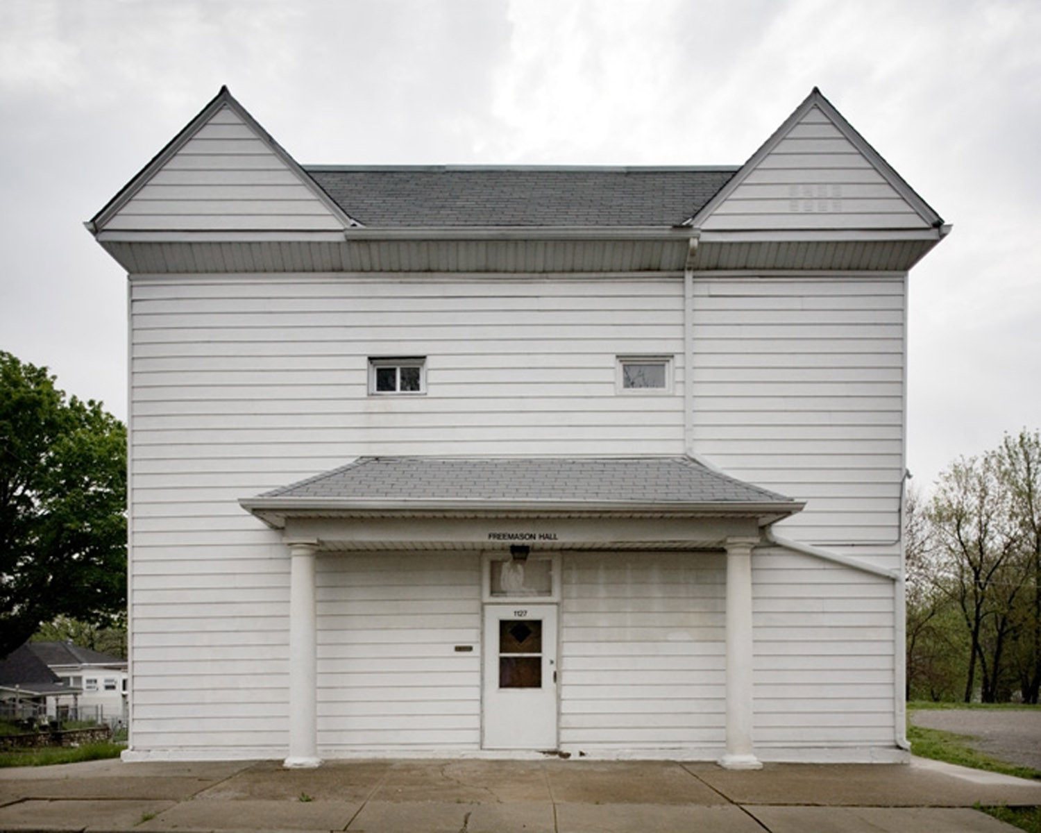 A symmetrical frontal view of a two-story white wooden building with two peaked gable roofs and a centered portico supported by classical columns. Horizontal siding covers the facade, with small rectangular windows on the upper story and a dark wooden door beneath an overhang at street level.