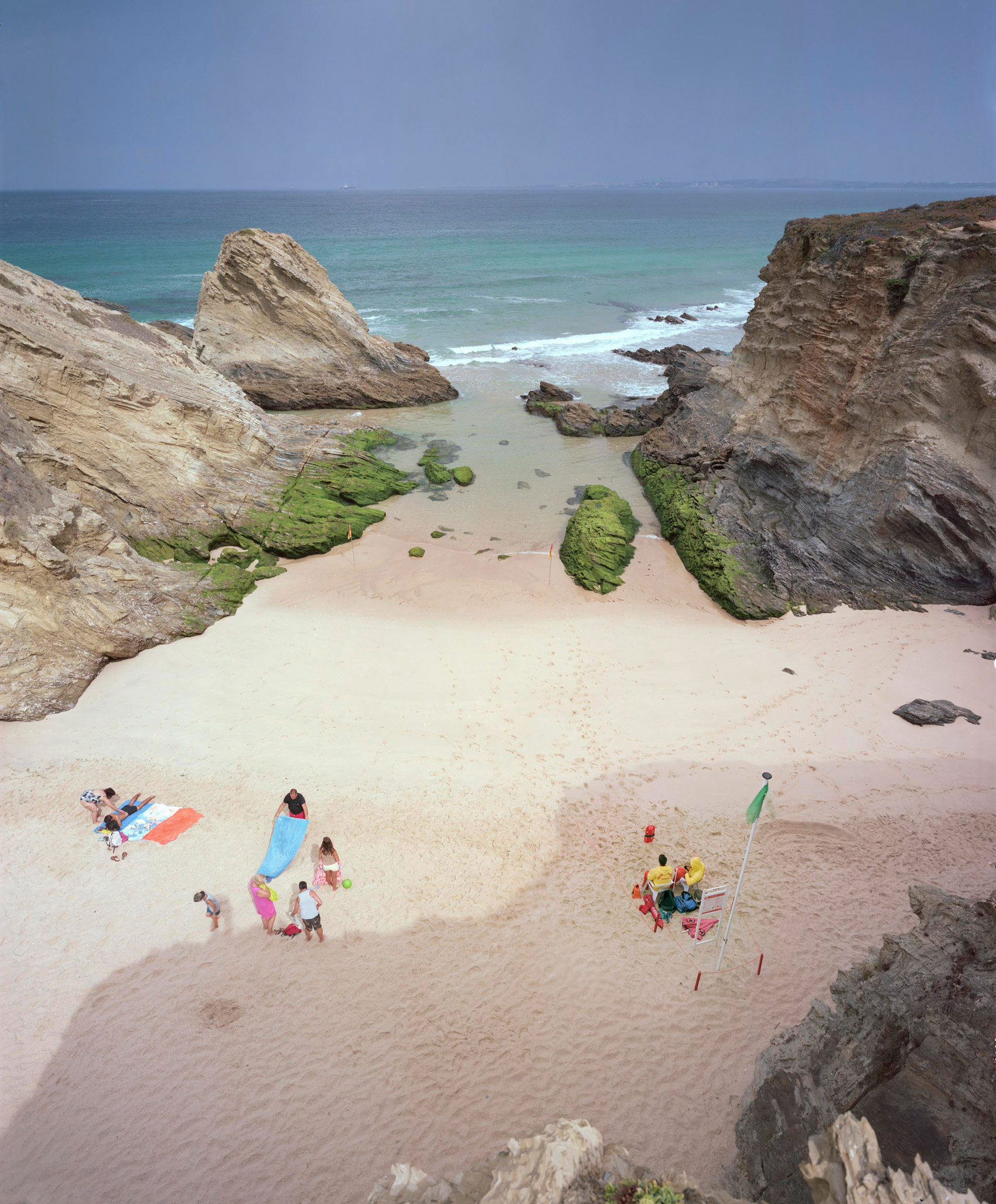 Aerial view of a secluded beach cove framed by dramatic layered rock formations, with turquoise water and white sand. Small groups of beachgoers are scattered across the shore, some with colorful towels and beach equipment, while moss-covered rocks and boulders dot the sandy beach and shallow waters.