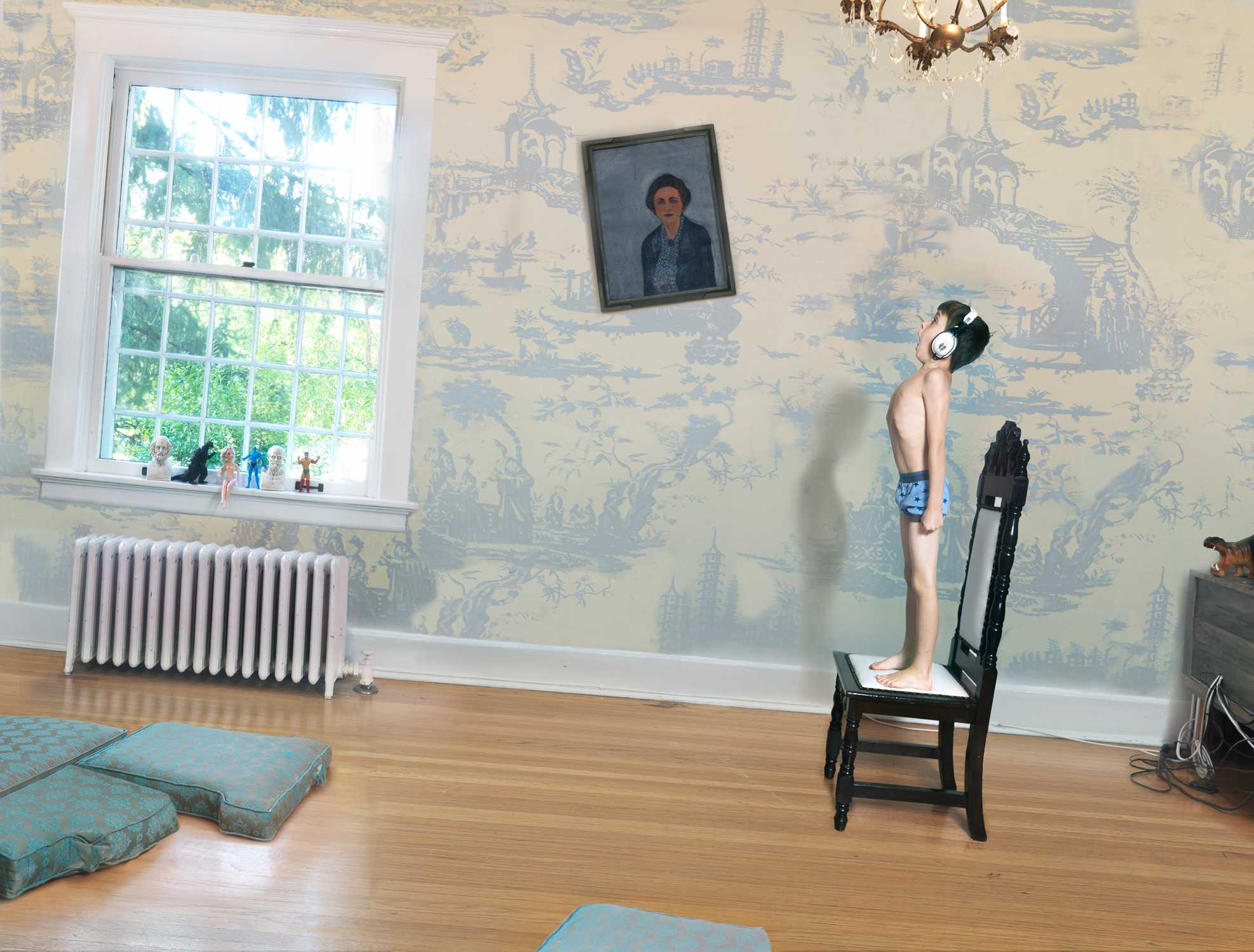 A shirtless boy wearing headphones stands on a black chair in a domestic interior, gazing upward. The room features cream and blue toile wallpaper, a radiator beneath a multi-paned window with small figurines on the sill, a portrait hanging on the wall, and blue cushions scattered on the wooden floor.