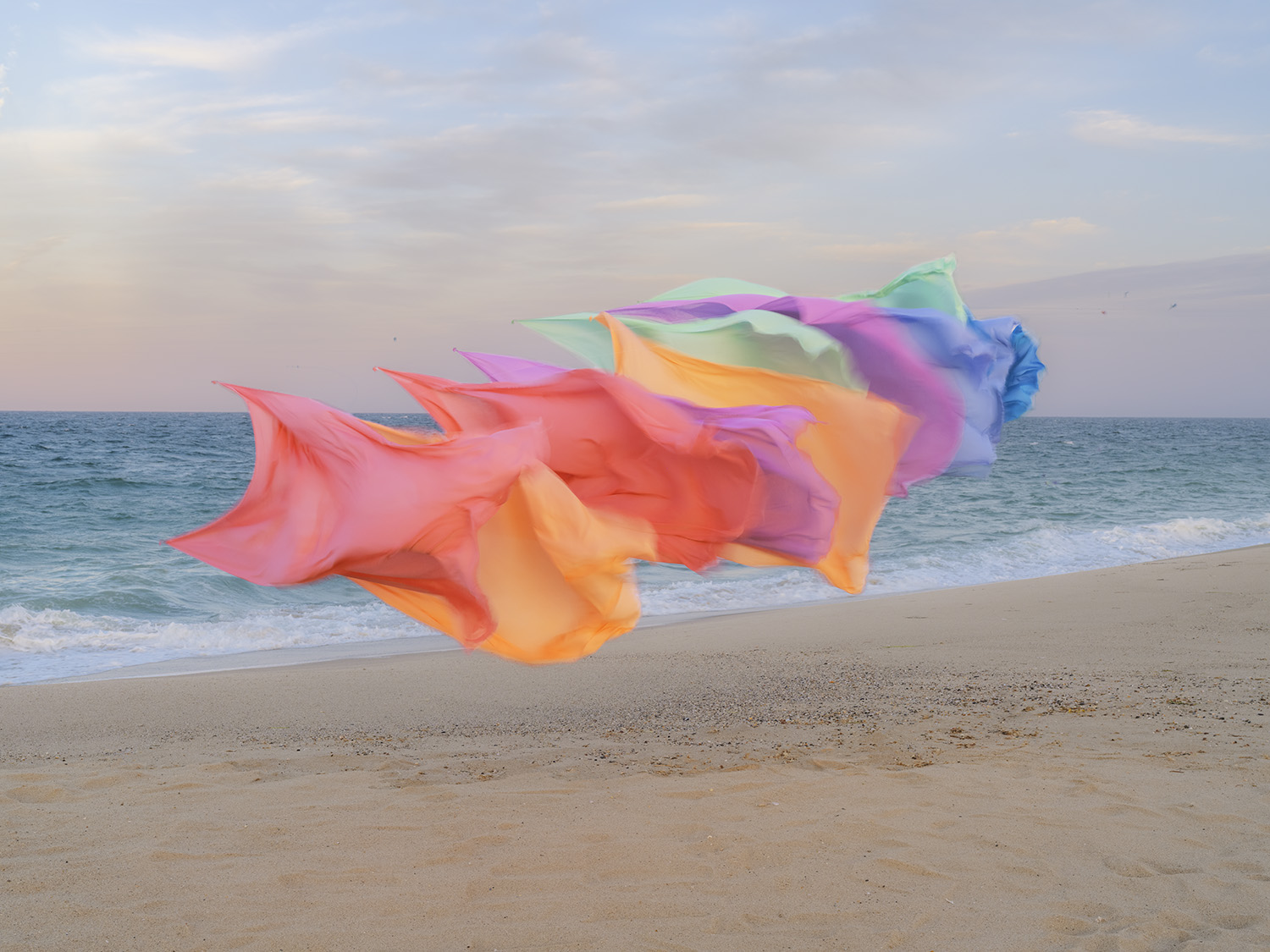 Billowing sheets of translucent fabric in gradient hues of coral, pink, orange, yellow, purple, and blue float above a sandy beach, with calm ocean waves and overcast sky visible in the background.