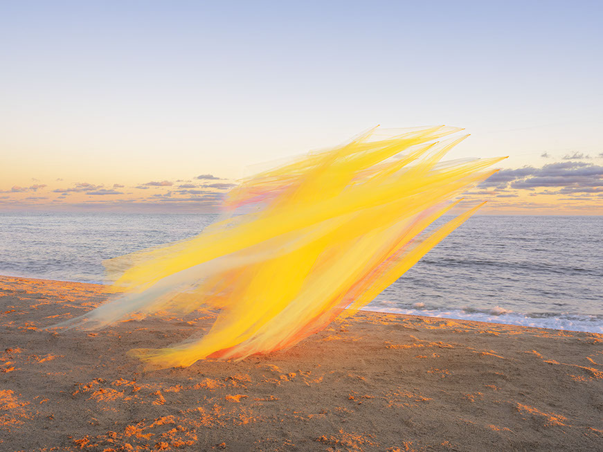 Billowing yellow and orange tulle fabric sweeps across a sandy beach at sunset, with the ocean and twilight sky visible in the background. The translucent, flowing material creates dynamic diagonal lines that contrast with the warm sand and cool water below.