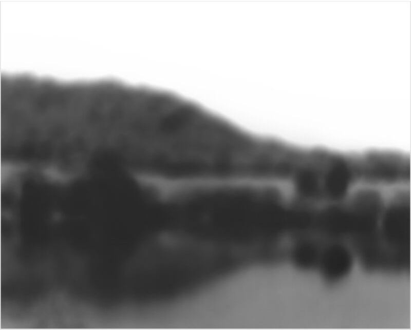 Softly focused black and white landscape depicting a body of water with forested hills or mountains rising beyond it, rendered in subtle tonal gradations that emphasize atmospheric depth and the ephemeral quality of the scene.