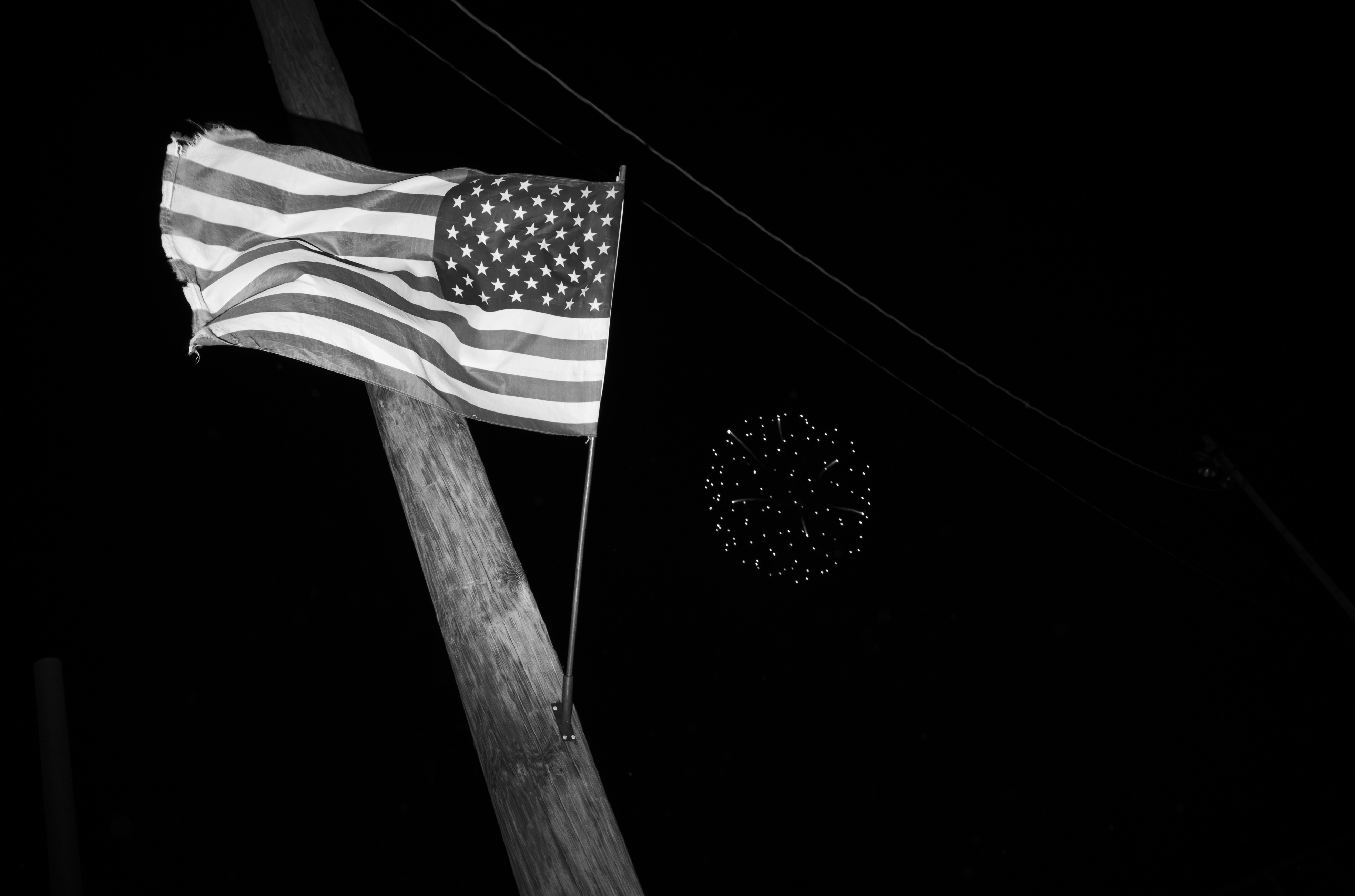 American flag mounted on a wooden pole against a black night sky, with fireworks bursting in the distance. The black and white photograph captures the flag in sharp detail while the distant explosions appear as scattered points of light.