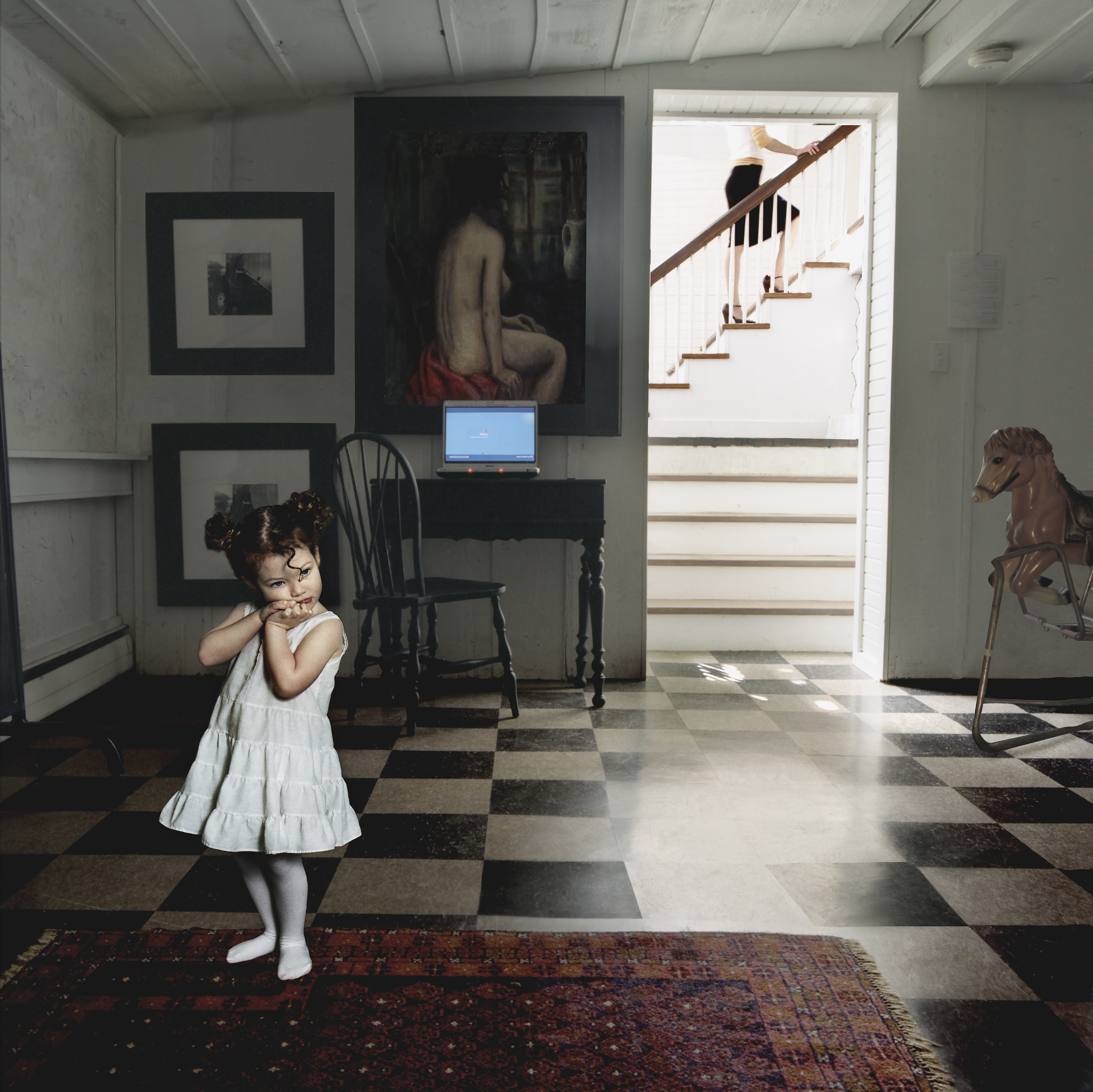 A young child in a white dress stands on a checkered floor in a bright, austere interior space, touching her neck with a contemplative gesture. The room features classical artwork on the walls, a black desk with a laptop, wooden stairs bathed in natural light, and a wooden rocking horse, creating a composed domestic scene with sharp contrasts between light and shadow.