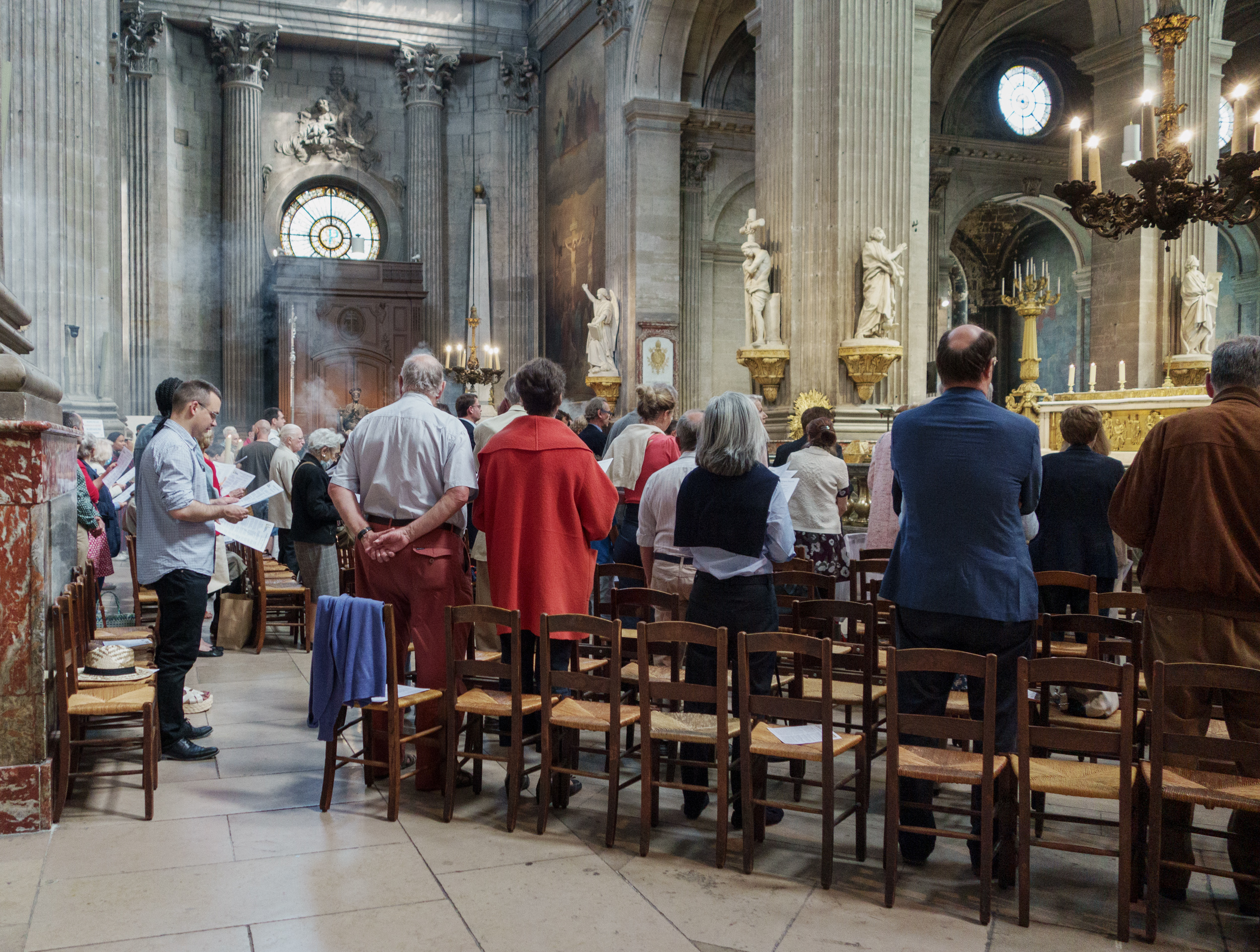 Congregation of visitors stands in the nave of a grand cathedral, facing toward the altar. Ornate architectural elements including soaring fluted columns, classical statuary on gilt pedestals, and a large circular clock window dominate the space, while wooden chairs are arranged throughout the foreground.