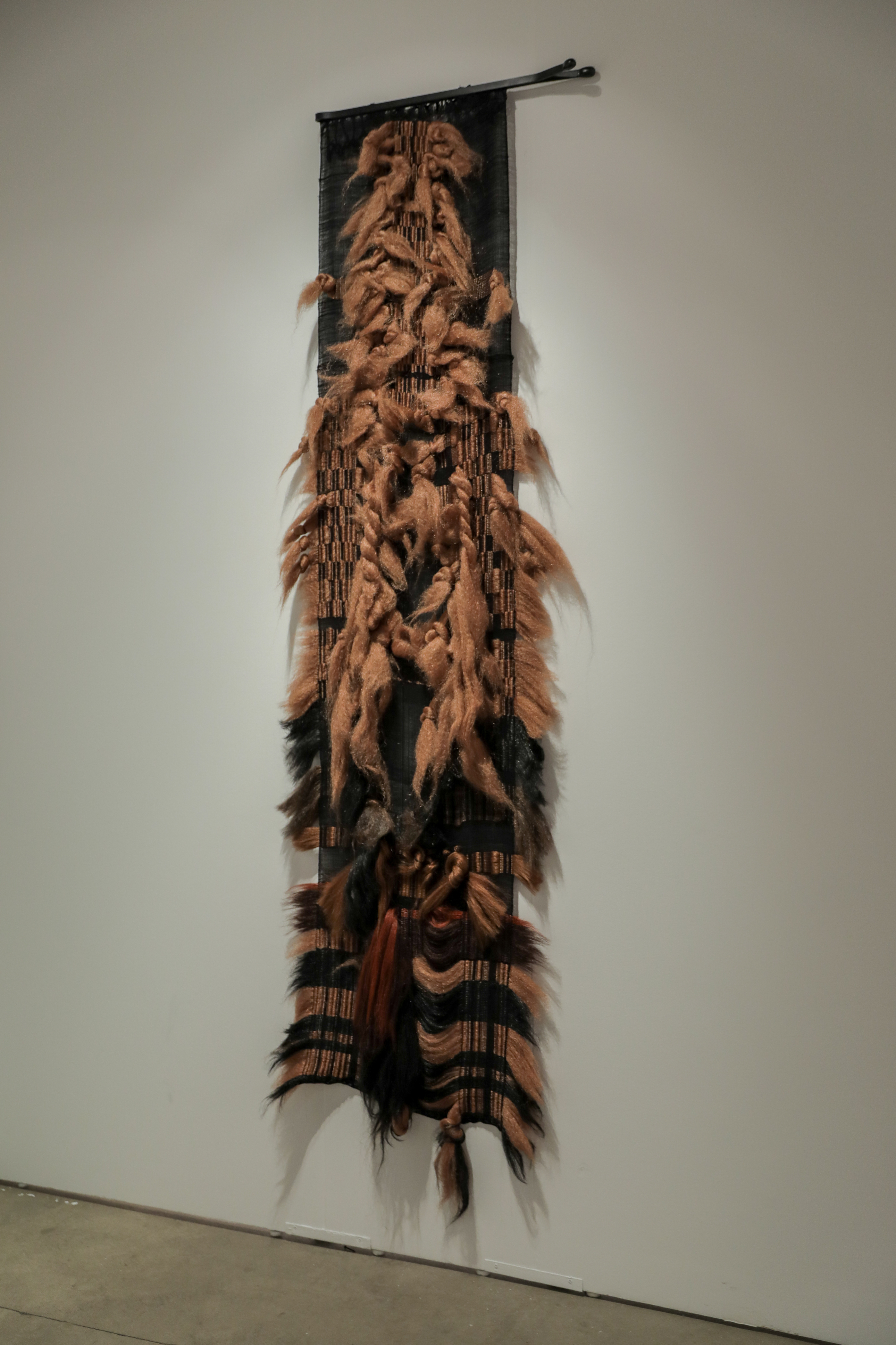 A vertical tapestry featuring expressive carved or sculpted faces in warm brown tones mounted on dark fabric, with synthetic hair and natural fibers extending outward along the sides, creating a textured, three-dimensional effect against a white gallery wall.