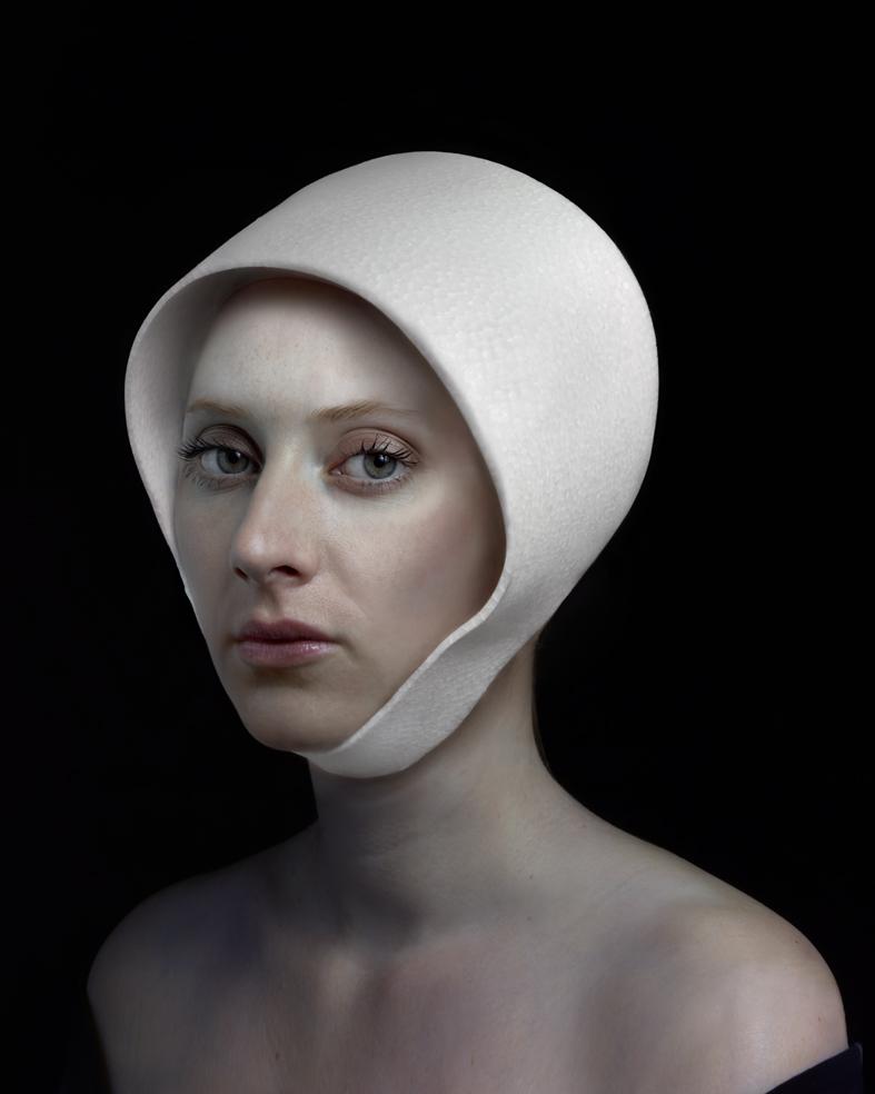 Portrait of a pale-skinned woman with dark eyes and lips against a black background, her head wrapped in a white fabric hood that frames her face. The minimalist composition emphasizes the subject's contemplative expression and the stark contrast between her skin tone and the pristine white textile.
