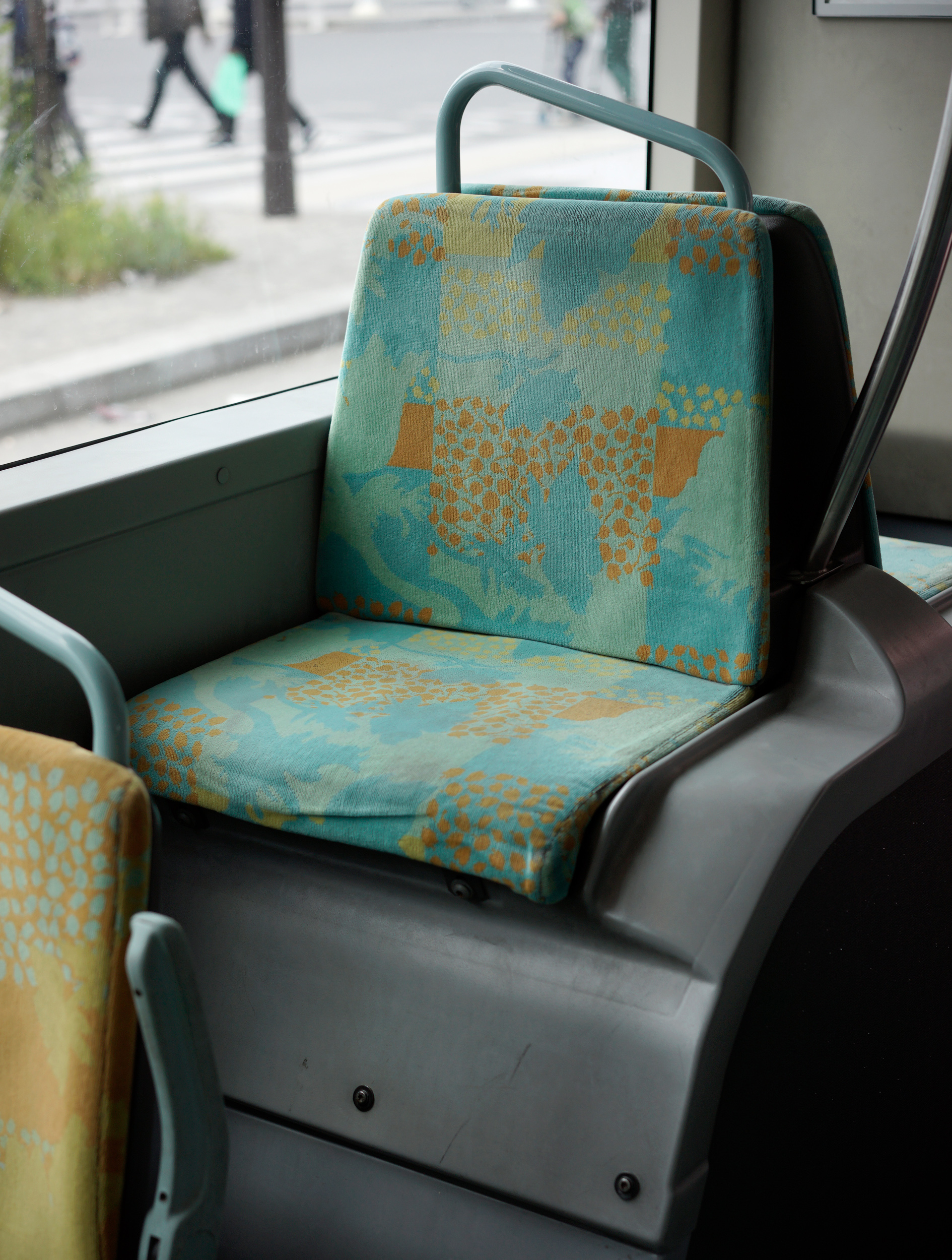 Public transit seat with turquoise and ochre patterned fabric upholstery featuring abstract geometric motifs, photographed from inside a bus or tram with an urban street scene visible through the window behind.