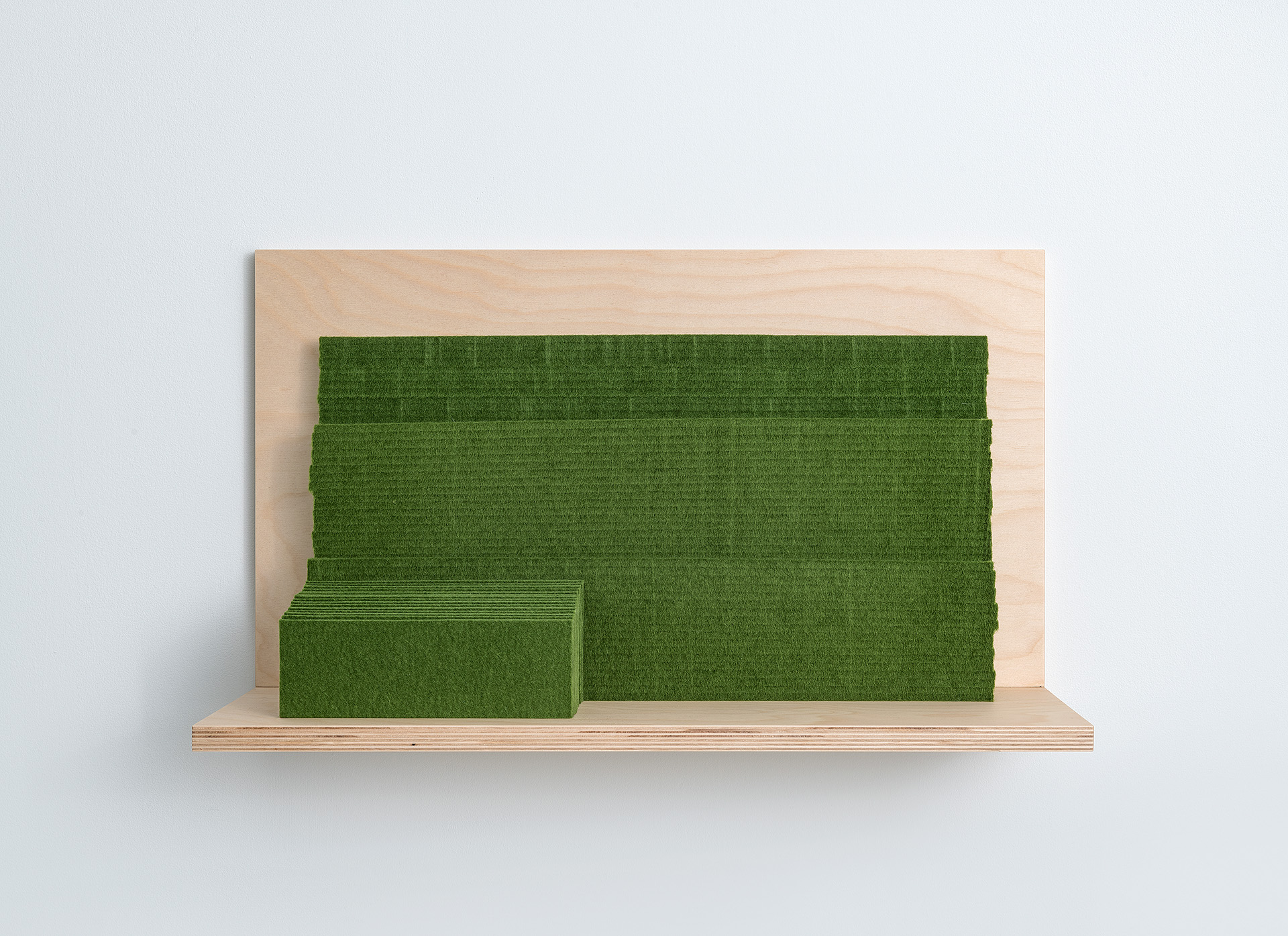 A wooden shelf mounted on a white wall holds two green felt rectangular forms of different sizes—a smaller one positioned on the left and a larger one behind it—creating a minimalist sculptural composition.