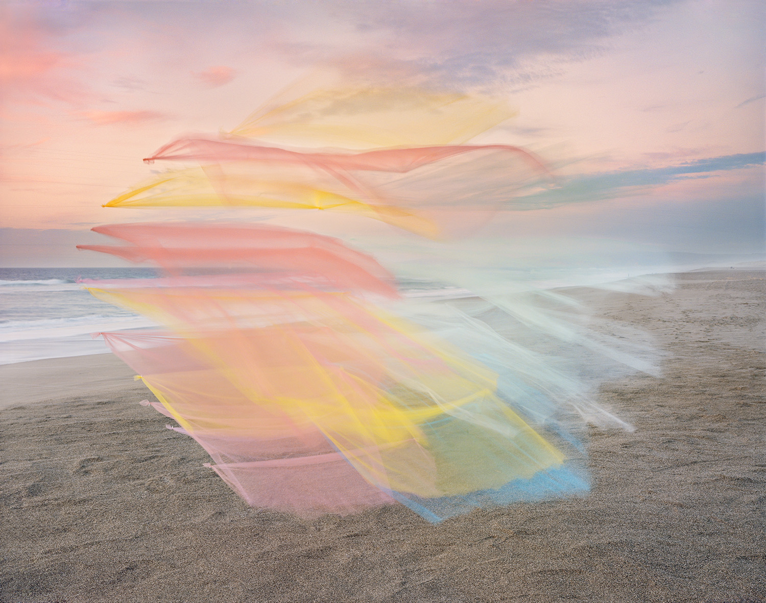 Layered translucent colored fabrics in shades of pink, yellow, and blue sweep across a sandy beach at twilight, creating an ethereal, flowing composition against the calm ocean and soft sky beyond.