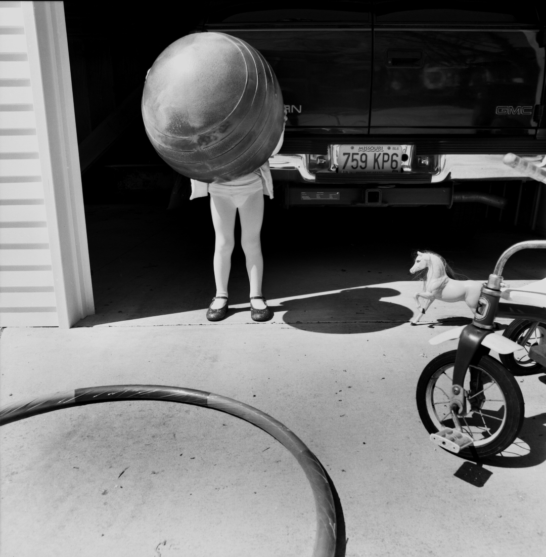 Black and white photograph of a person holding an enormous striped ball that obscures their head and upper body, standing in a driveway beside a GMC truck with visible license plate and a child's tricycle. A small toy horse figure sits nearby on the concrete pavement.
