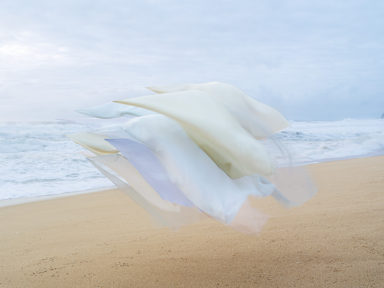 White and cream-colored silk fabric billows and folds across a sandy beach, with ocean waves and overcast sky visible in the background. The translucent material catches subtle shadows of blue and lavender as it moves in the wind.