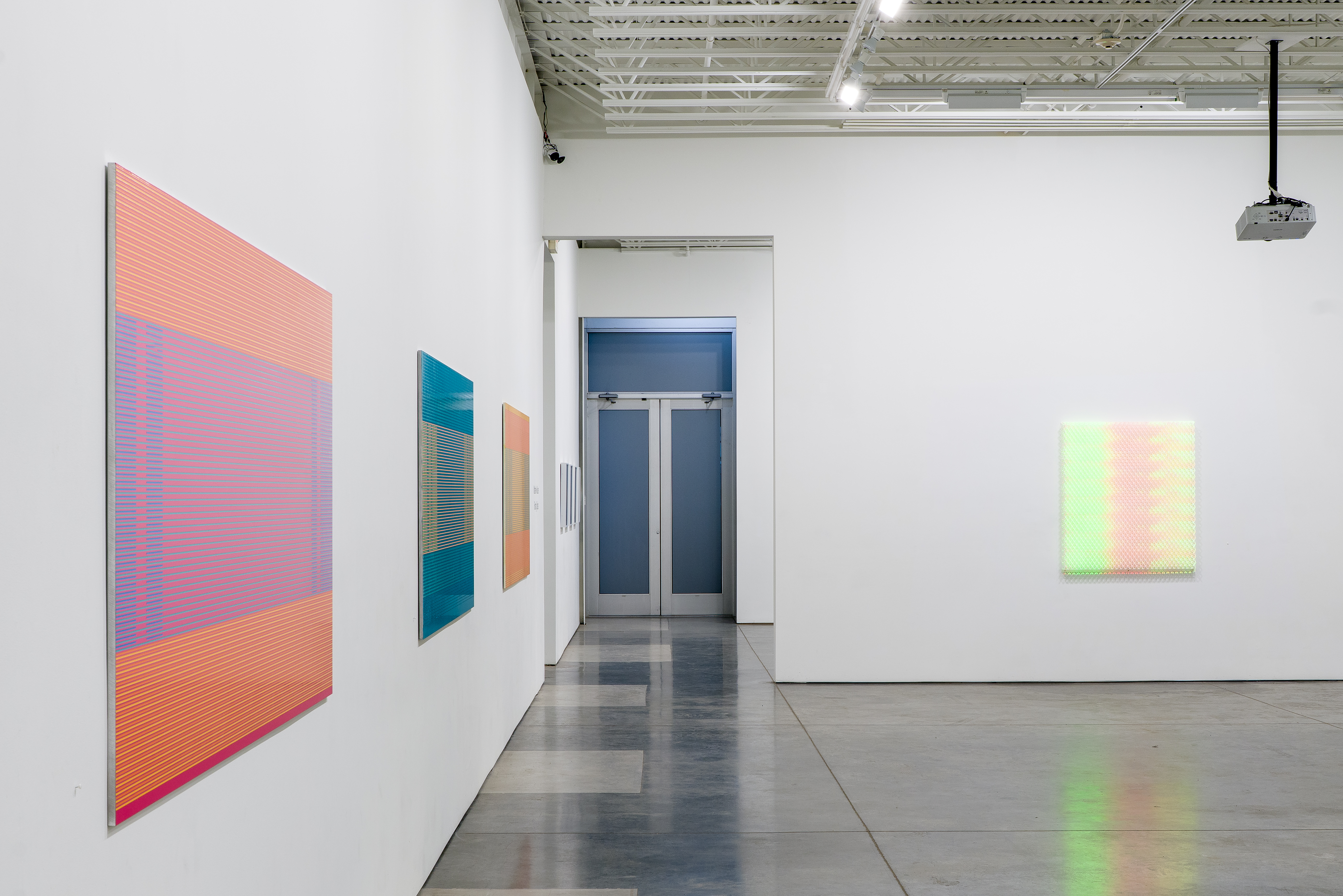 Contemporary gallery installation featuring large-scale abstract paintings with horizontal striped patterns in vibrant coral, pink, teal, and yellow hues displayed on white walls. Digital video projections cast colorful light onto select works, creating hybrid physical-digital compositions. The minimalist space includes polished concrete flooring and a blue doorway visible in the background.
