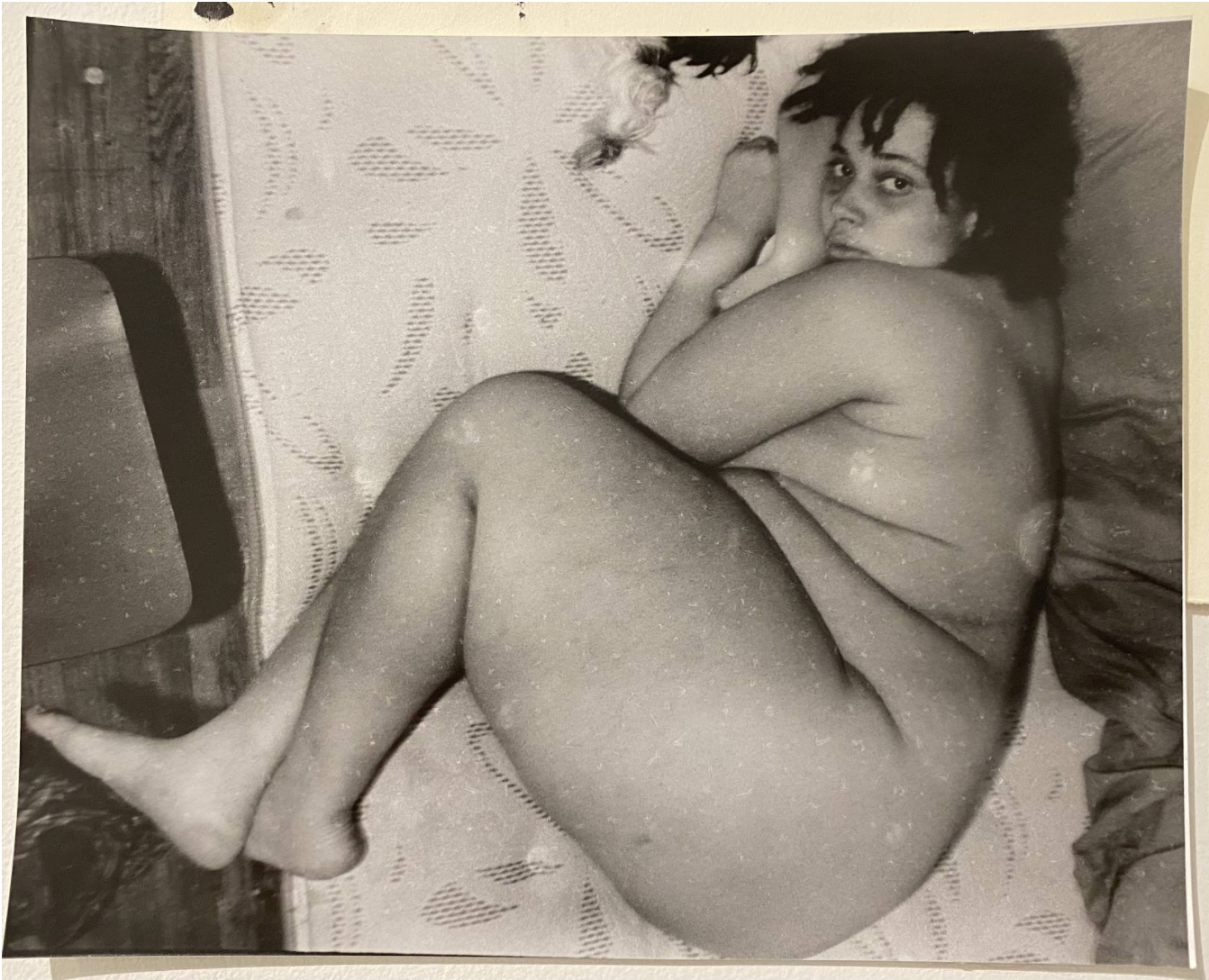 A black and white photograph depicting a reclining figure partially draped in textured bedding, shot from above at an intimate angle. The composition emphasizes the human form against patterned fabric, with dramatic lighting characteristic of silver gelatin printing.
