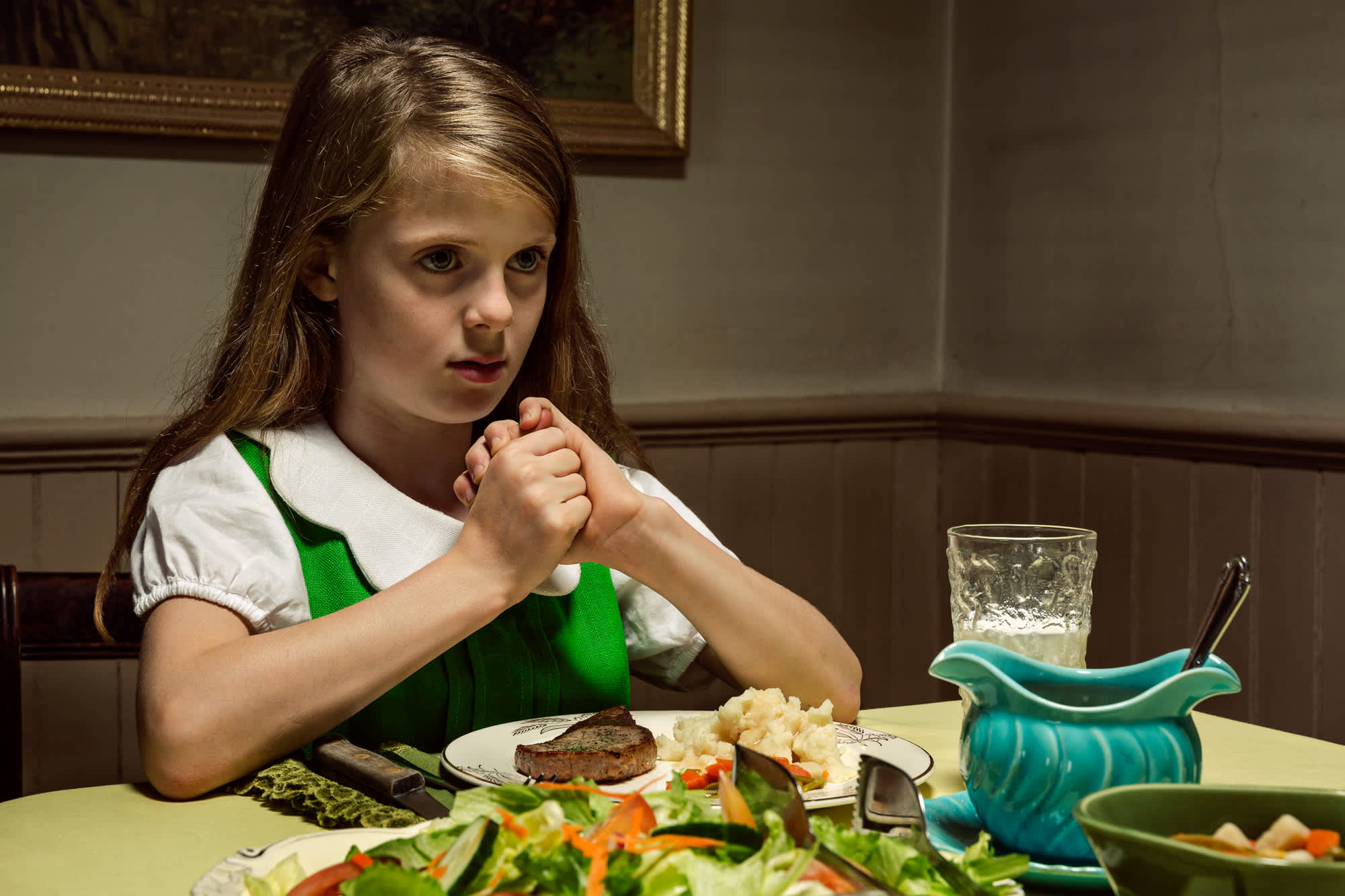 A young girl in a green and white dress sits at a dining table with her hands clasped, gazing downward at a plate of food before her. The table is set with various dishes including salad, meat, and potatoes, alongside a glass of water and turquoise ceramic serving vessels, creating a formal domestic scene rendered in rich, naturalistic color.