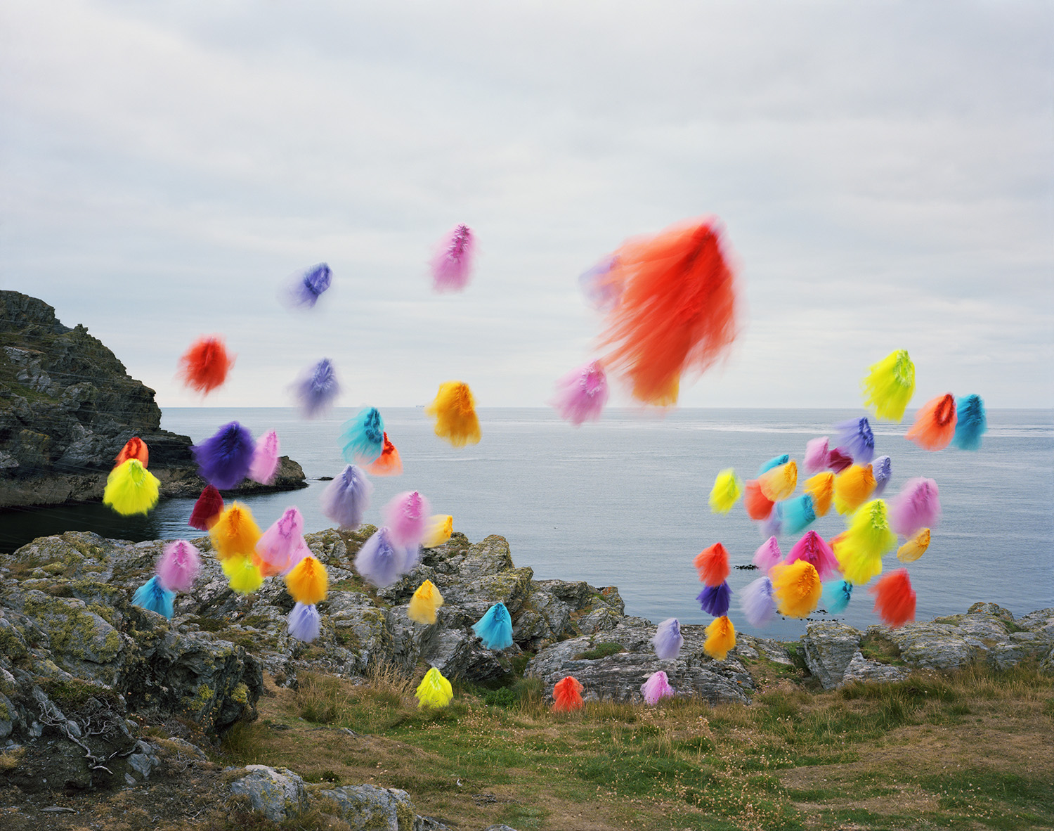 Vibrant colored tulle tutus in shades of red, pink, yellow, blue, orange, and purple are suspended mid-air and arranged across rocky coastal terrain overlooking calm seawater, creating a surreal contrast between the delicate fabric forms and the stark natural landscape.