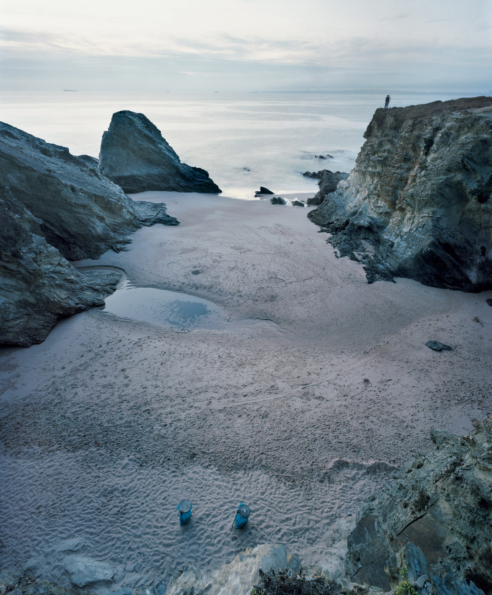 A coastal landscape at dusk featuring a sandy beach bordered by dramatic grey rock formations and sea stacks. Two small blue objects sit on the wet sand in the foreground, while a solitary figure stands on a distant cliff to the right. The calm, pale waters and overcast sky create a cool, muted color palette of greys, blues, and whites.