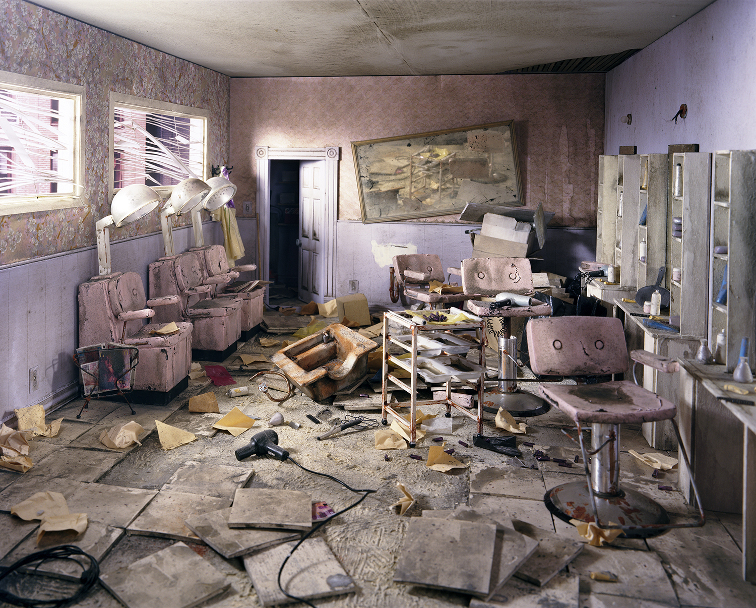 An abandoned interior of a dilapidated beauty salon with mauve-painted walls features overturned pink upholstered chairs, scattered debris and flooring materials, vintage salon equipment, and a faded landscape painting, rendered in vivid color despite the deteriorated state of the space.
