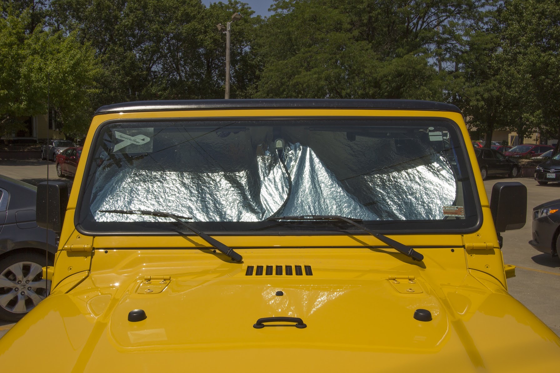 Bright yellow Jeep photographed from the front, its windshield covered with a reflective silver sunshade. The vehicle is parked in a lot with trees and other cars visible in the background, creating a stark contrast between the vivid yellow body and the metallic shimmer of the protective shade.