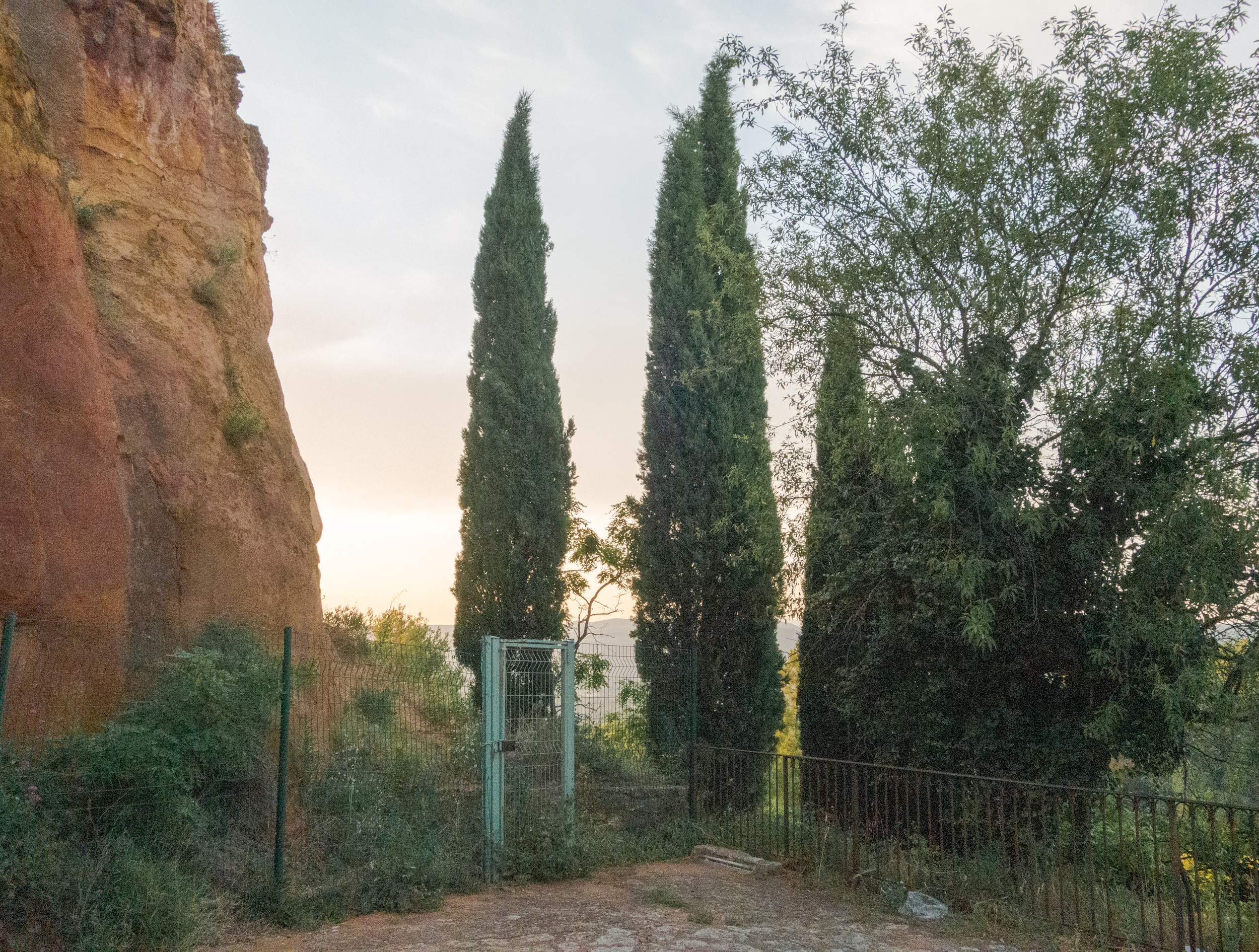 A rustic stone fortification wall rises on the left beside a narrow dirt path bordered by metal fencing. Three tall, narrow cypress trees stand prominently in the center, flanked by lighter deciduous foliage on the right. A turquoise metal gate marks an opening in the path ahead, with a distant landscape visible beneath a soft, overcast sky.