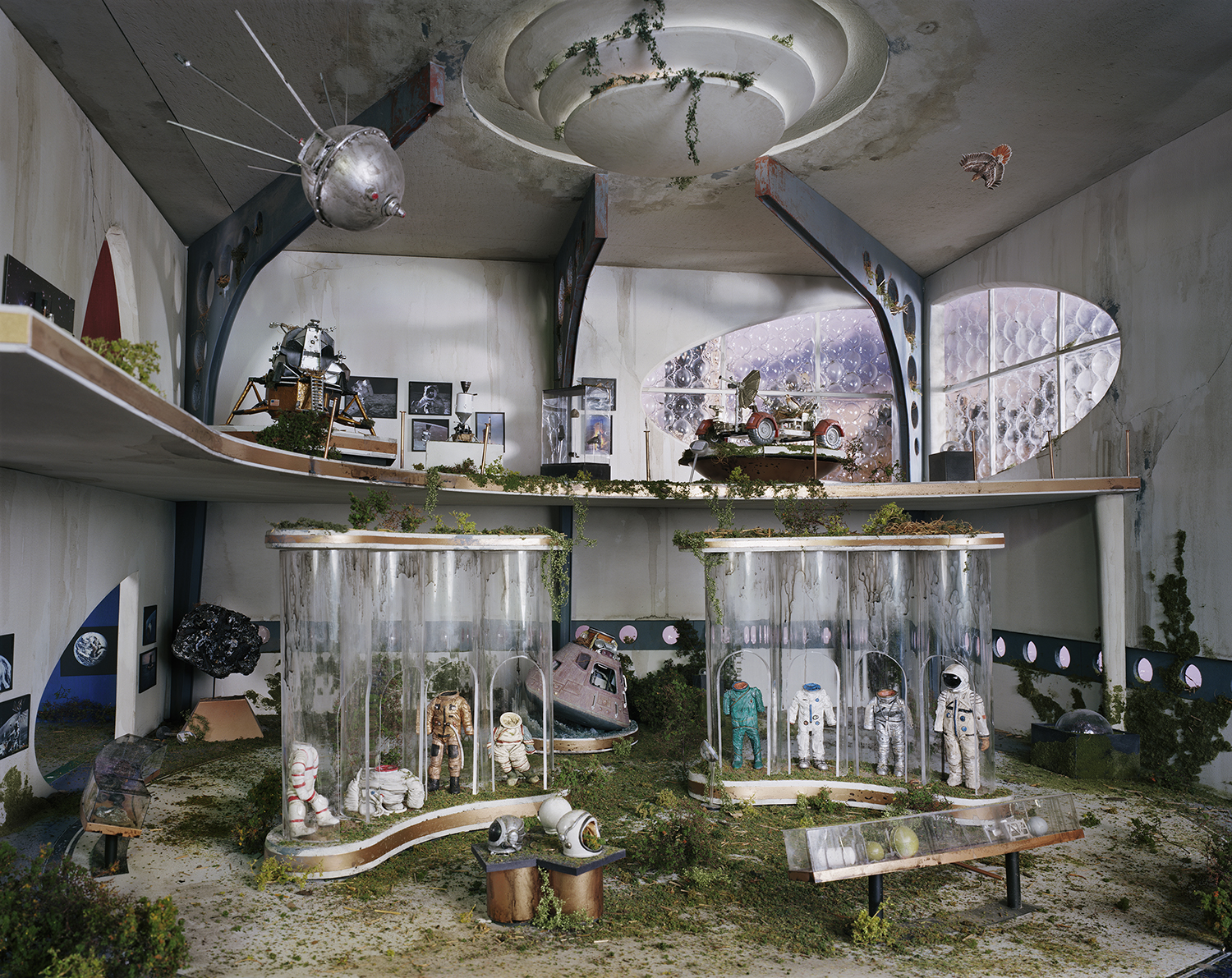 An abandoned institutional interior overgrown with vegetation features glass display cases containing astronaut suits and space equipment, with a Soyuz capsule and lunar rover visible on upper shelves. Concrete walls, debris-strewn floors, and celestial imagery suggest a decaying space museum frozen in time.