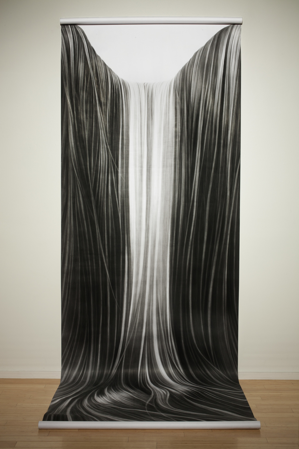A monumental vertical drawing in charcoal depicts flowing, hair-like strands cascading downward in sinuous curves. The composition features darker tones at the edges that gradually lighten toward a luminous central channel, creating a sense of movement and depth as the linear forms pool and gather at the base.