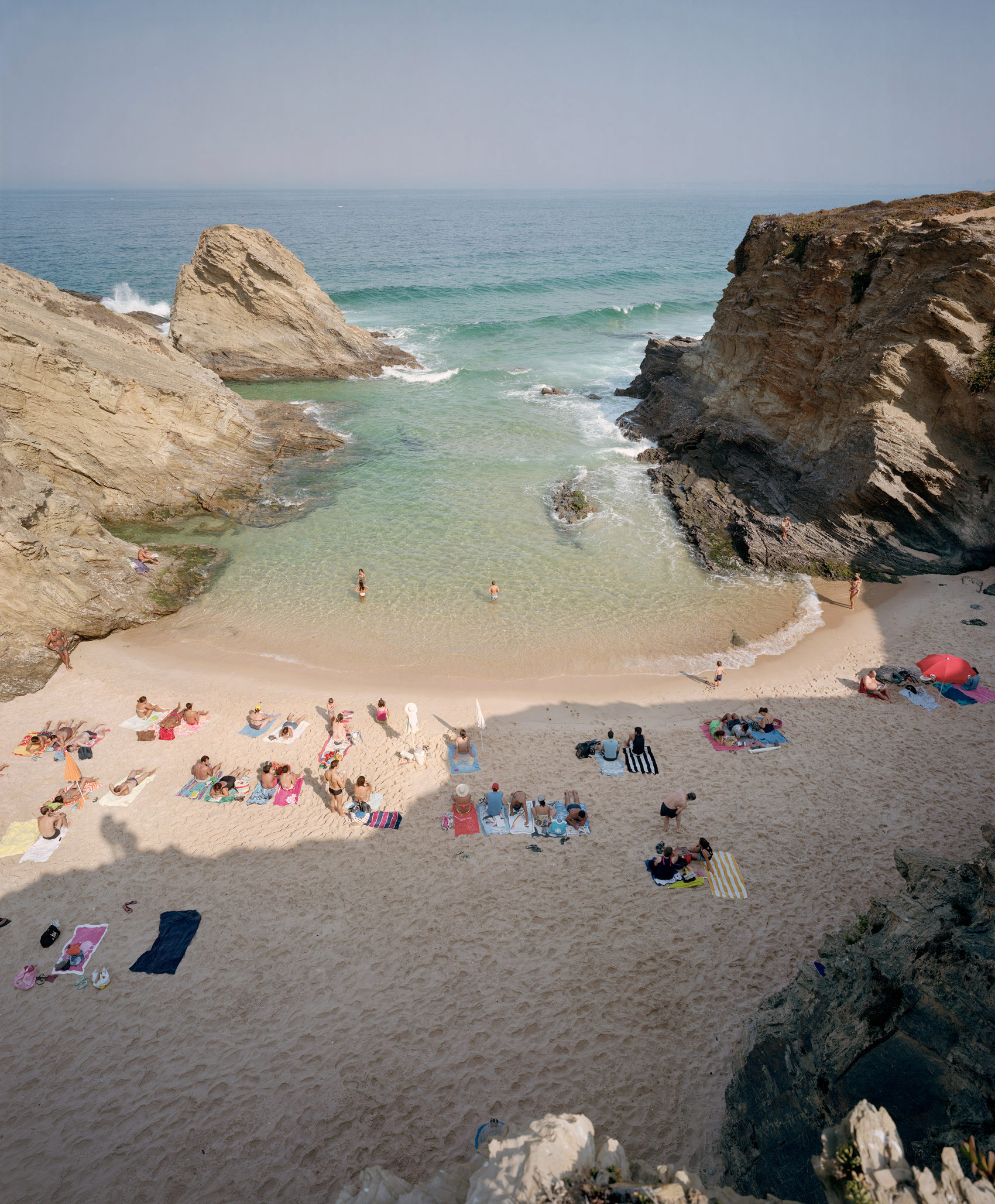 Aerial view of a secluded cove beach framed by dramatic limestone cliffs, with scattered beachgoers in colorful clothing and towels on pale sand, turquoise shallow waters, and rocky outcrops creating a sheltered bay.