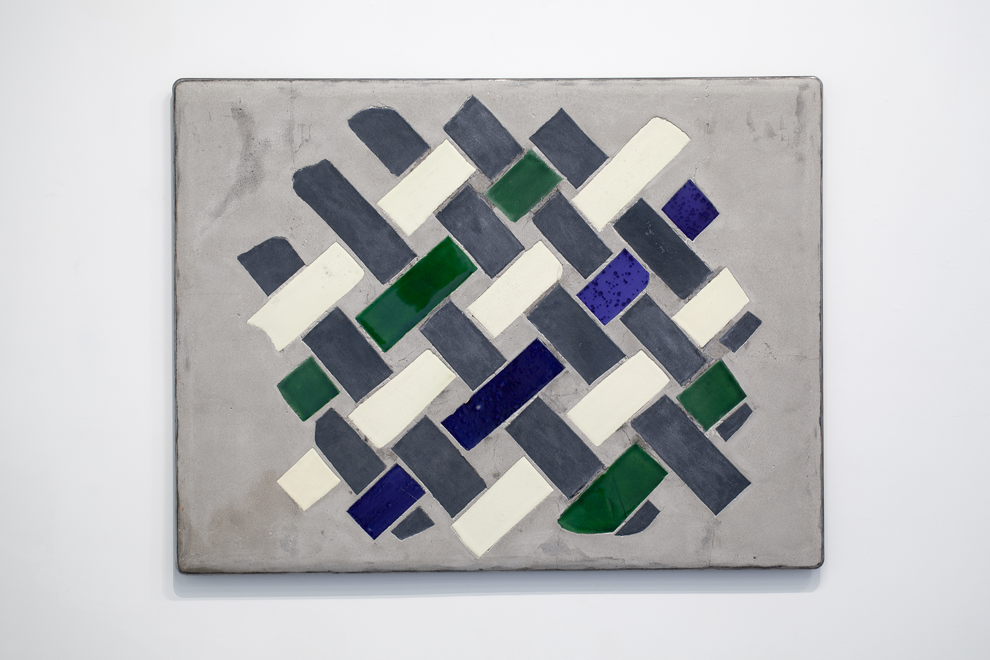 Concrete rectangular panel featuring a herringbone pattern of colored ceramic tiles in dark gray, cream, deep green, and cobalt blue, arranged diagonally across the surface with subtle shadows creating depth and dimension.