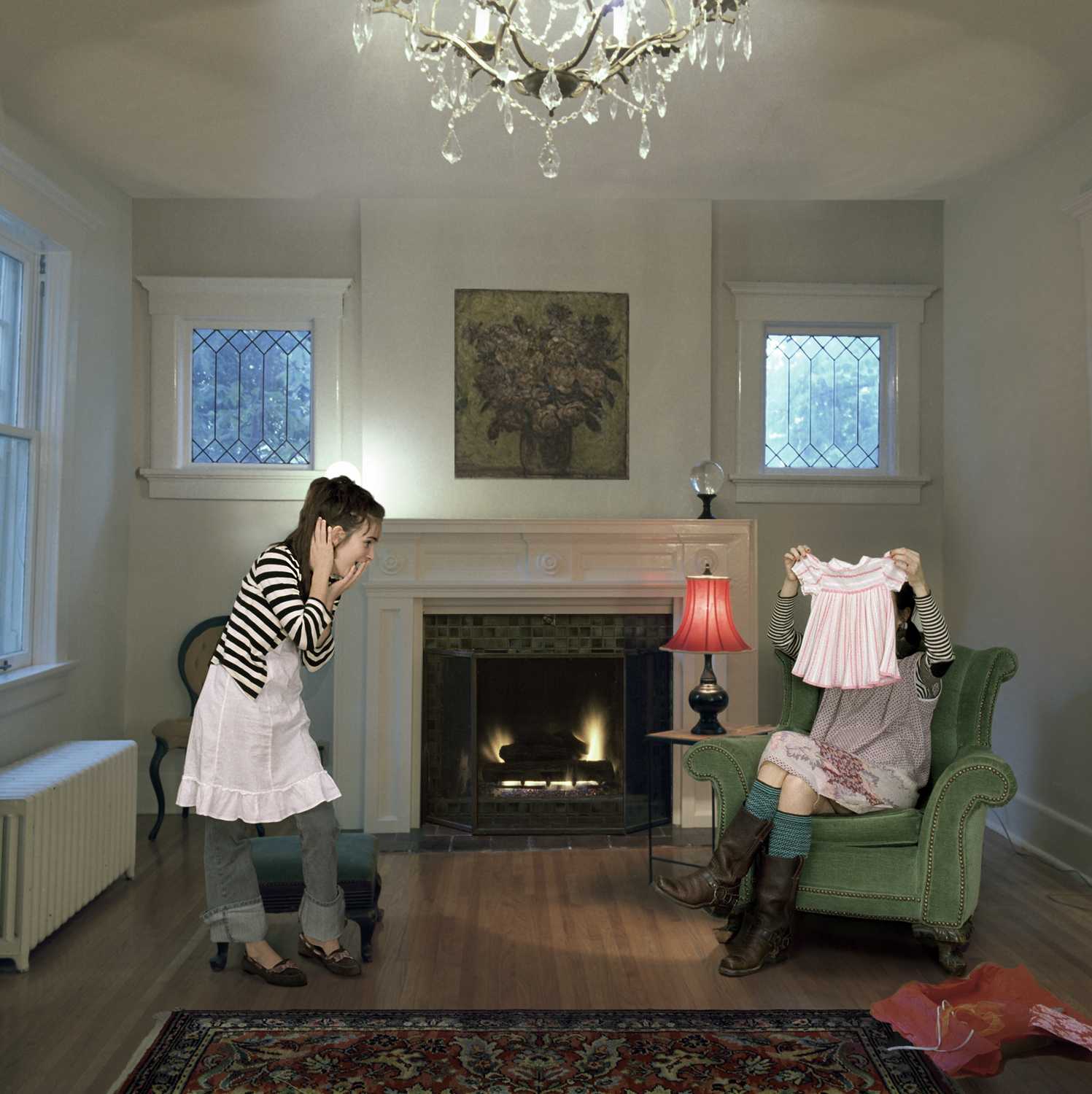 A woman in a striped shirt and white skirt stands in a elegantly appointed living room, holding up a pink children's dress toward a seated mannequin dressed in period clothing. Sunlight streams through leaded-glass windows flanking a fireplace with a red-shaded lamp, while a crystal chandelier hangs overhead and an ornate rug anchors the composition.