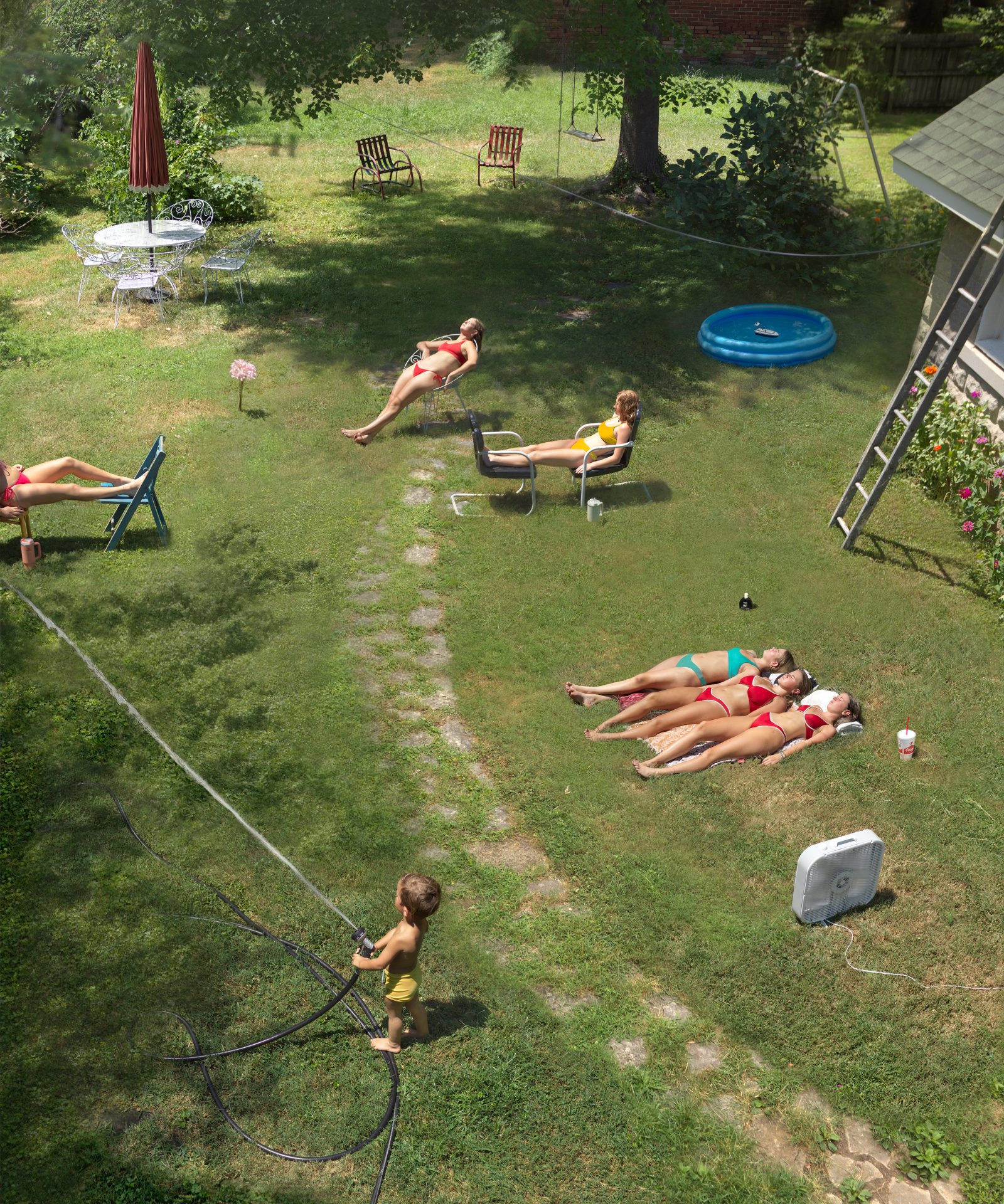 Aerial view of a suburban backyard where multiple people in swimwear are scattered across a lawn engaging in various leisure activities—some lying down, others sitting in chairs or doing exercises—surrounded by pool equipment, garden furniture, and a blue inflatable pool, creating a vibrant scene of summer relaxation.