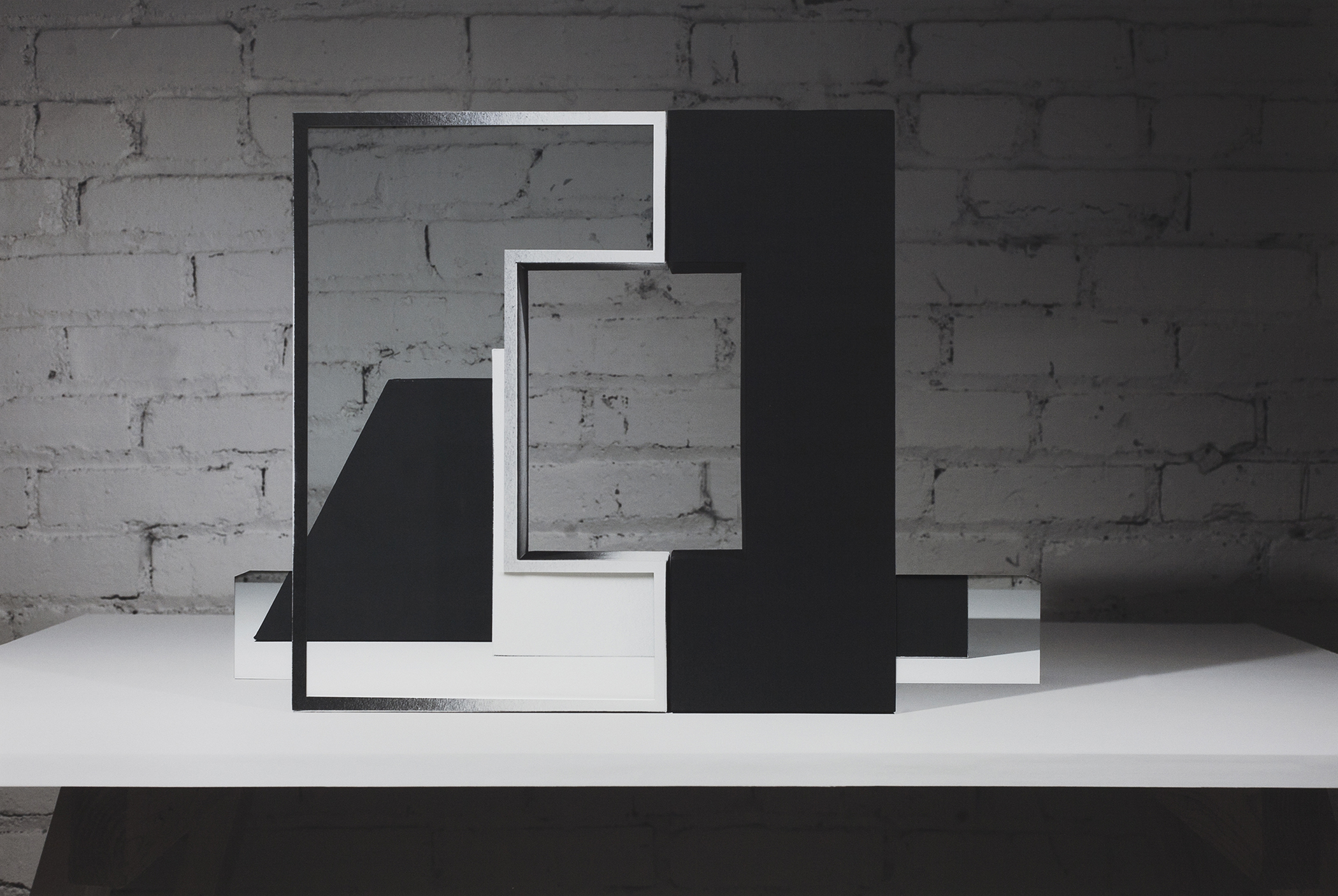 Abstract geometric composition of layered rectangular frames in black and white, creating a three-dimensional architectural form against a weathered brick wall. The overlapping planes and frames cast shadows on a white surface, establishing depth through contrasting solid and void spaces.