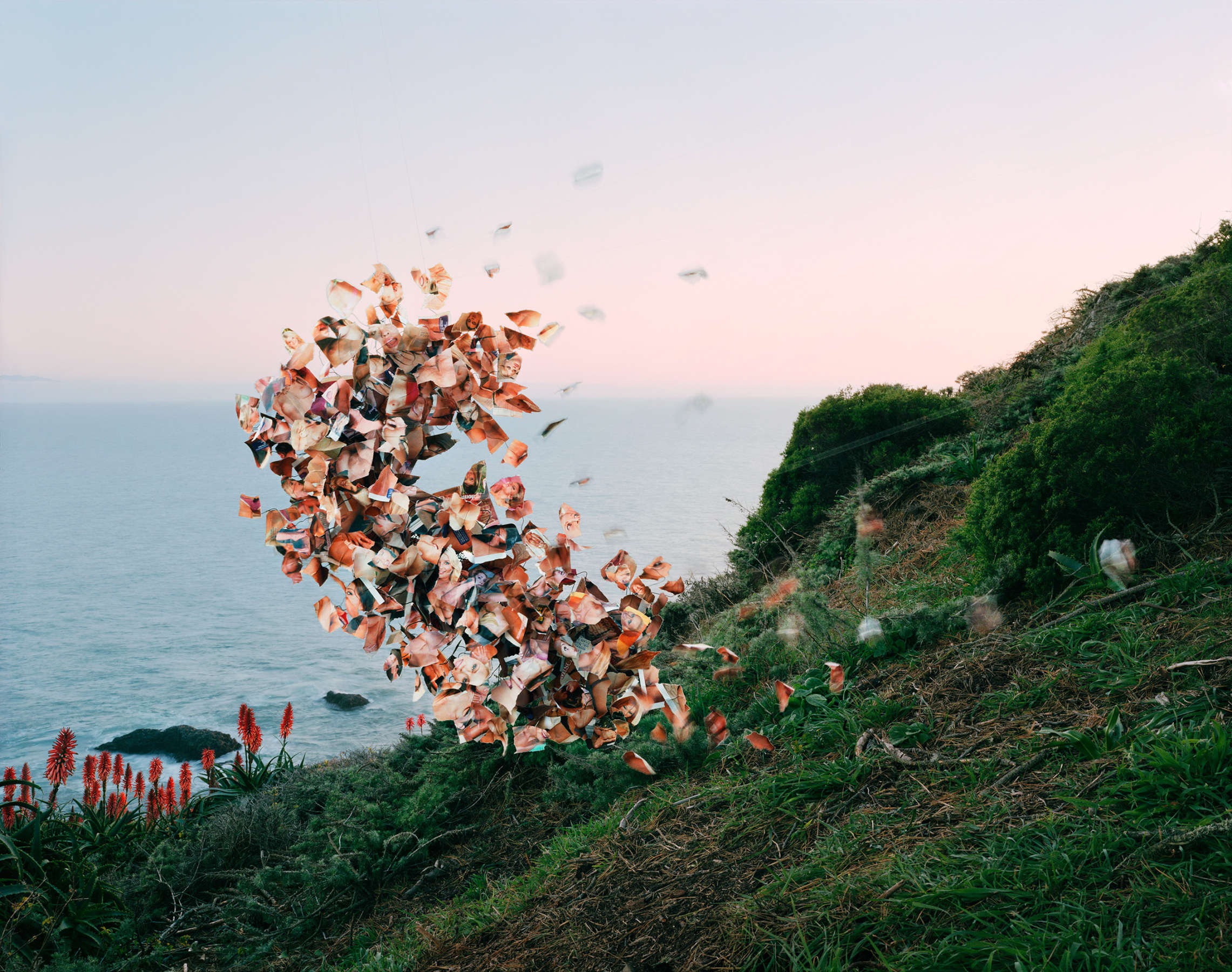A cluster of terracotta and copper-toned fragments suspended in mid-air above a coastal hillside, dispersing like an explosion or dissolution. Lush green vegetation and red flowering plants frame the foreground and right edge, while a calm sea and twilight sky occupy the background in soft blues and pinks.